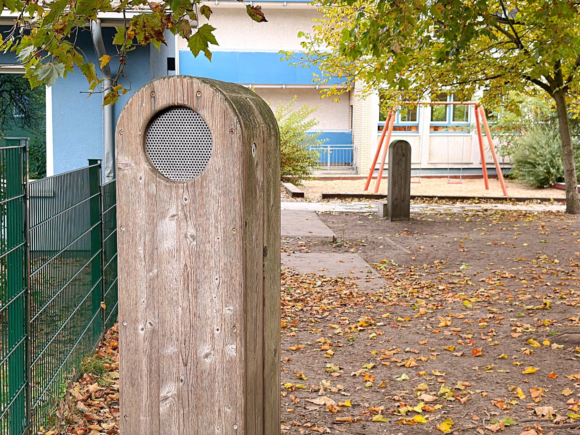 Blick in den Schulhof: im Vordergrund eine Stele aus Holz mit integriertem Lautsprecher, im Hintergrund eine Schaukel.