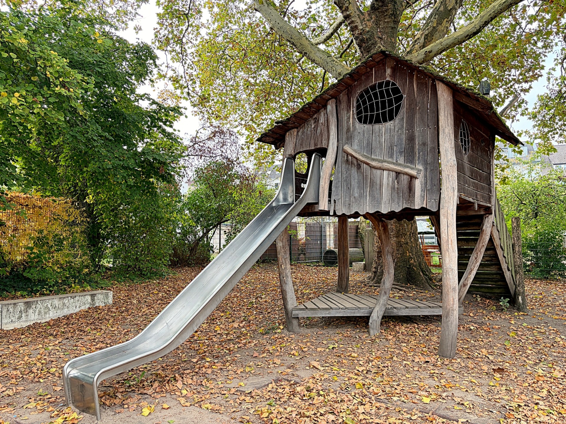 Ein großes Kletterhaus aus Holz, daraus führt eine silberne Rutsche.