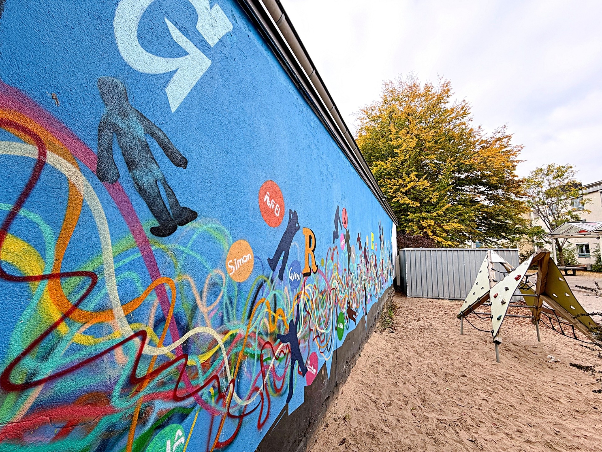 Eine blaue Wand mit einem bunten Graffittigemälde. Davor ist eine Sandfläche auf der ein Klettergerüst steht.