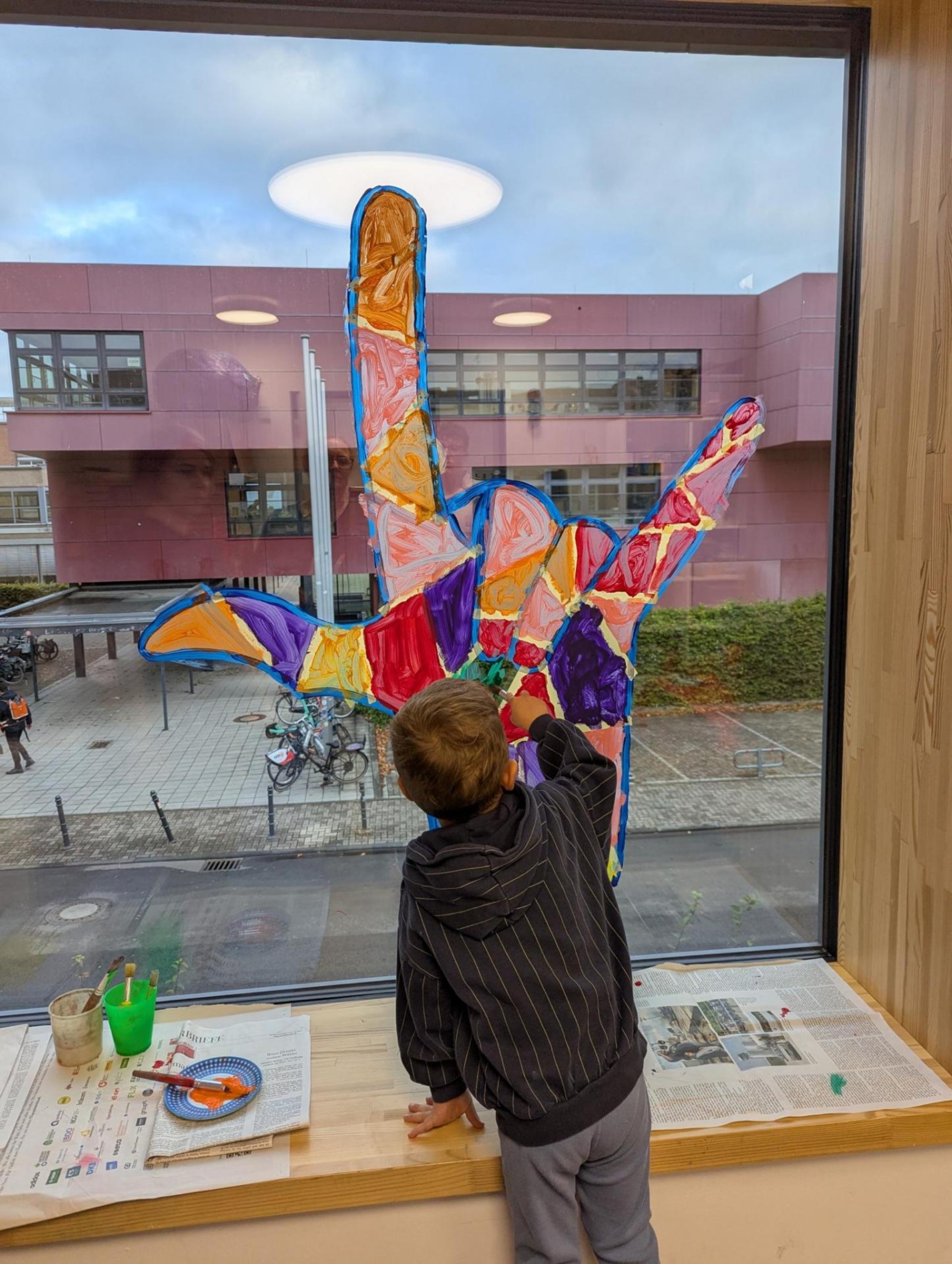 Ein Kind malt mit Fingerfarben eine sehr große, bunte ILY-Hand an einem Fenster aus.