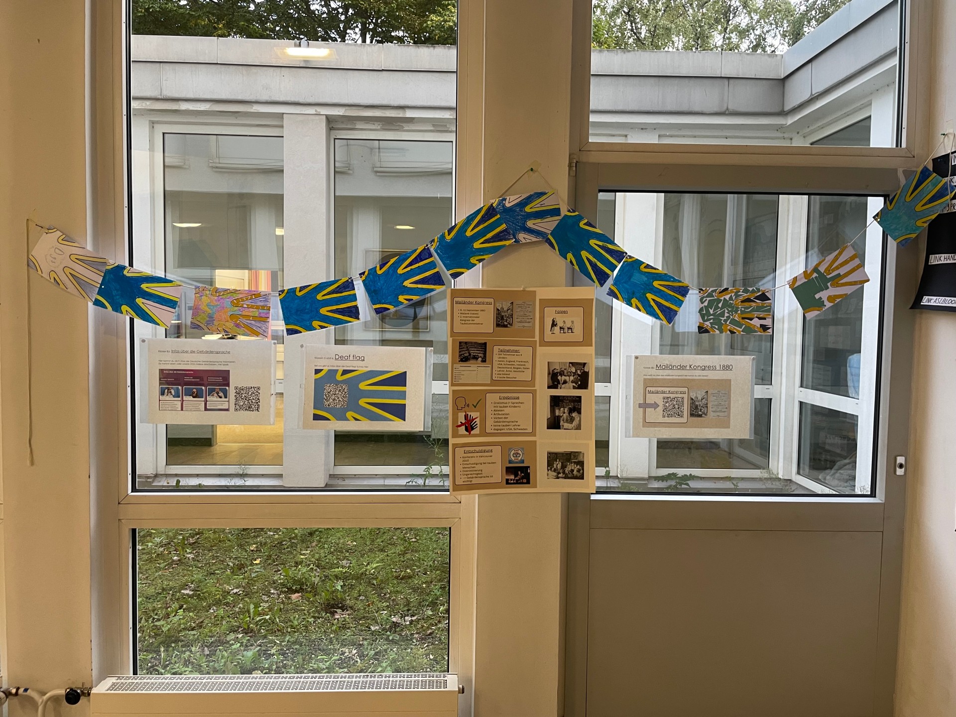 Vor einem Fenster hängen DIN A6-große Wimpel mit bunt gestalteten Gehörlosen-Flaggen und Infoplakate zu dieser Flagge.