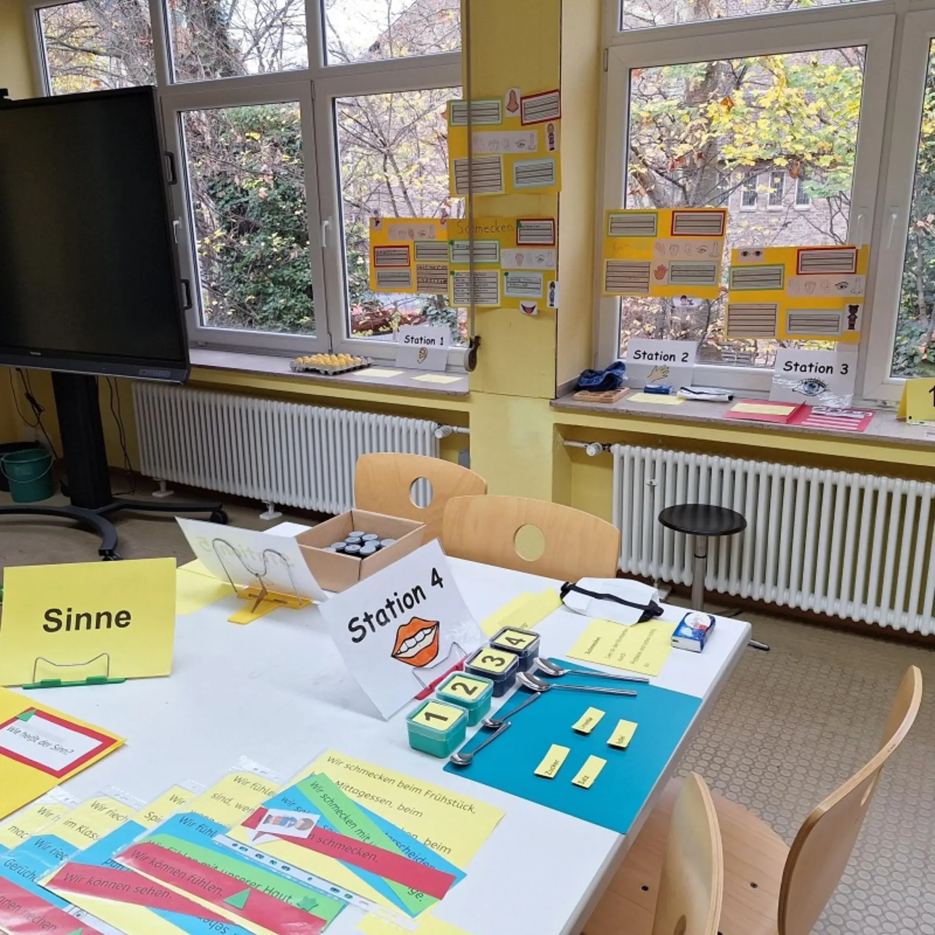 In einem Klassenraum steht ein Tisch mit Material zum Thema "Sinne", zum Beispiel kleine Dosen mit  den Zahlen 1-4 beschriftet und Löffel. An den Fenstern hängen Plakate zum Thema "Sinne". Auf dem Fensterbrett liegen weitere Materialien zu den Stationen 1, 2 und 3.