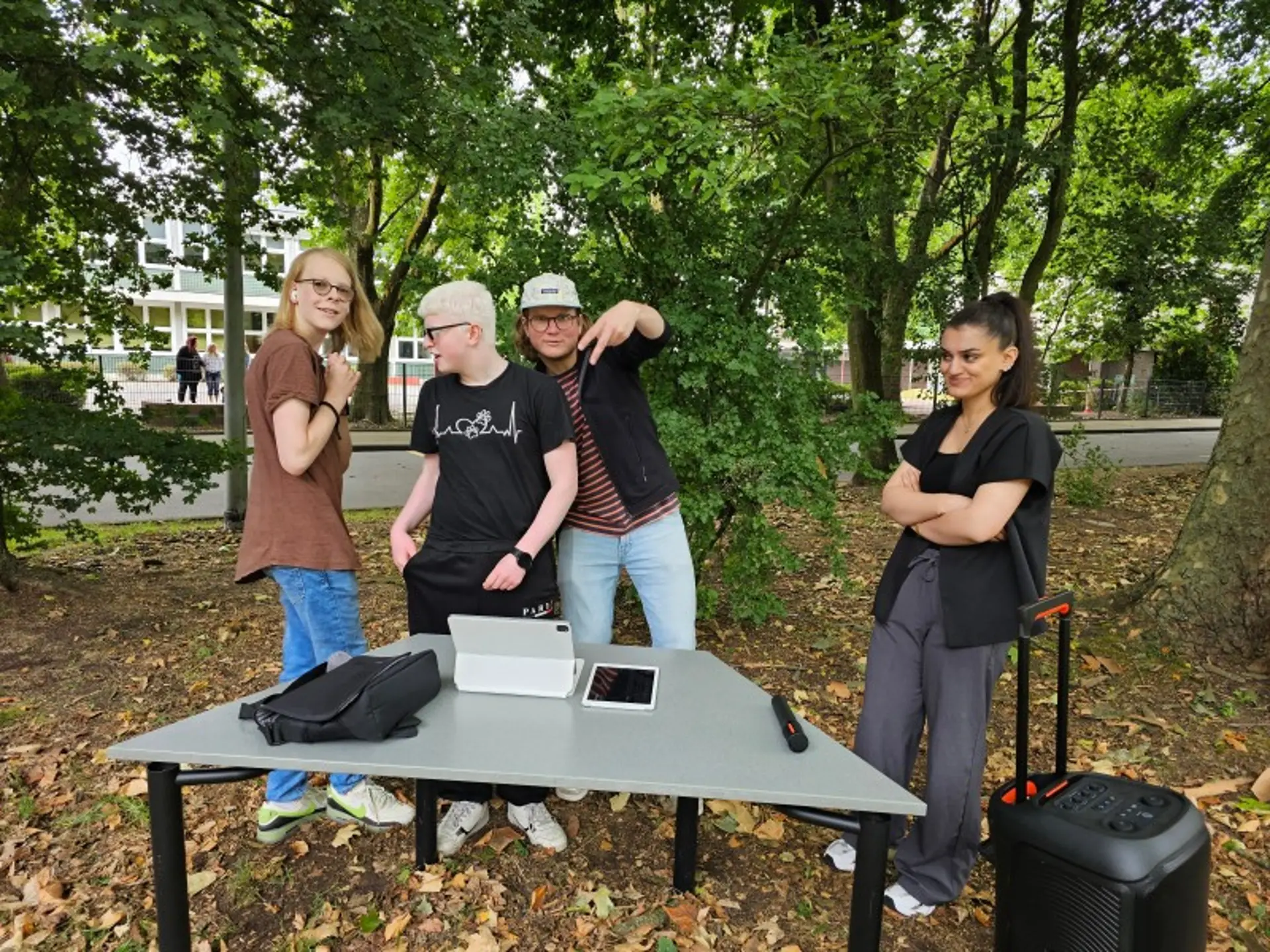 Vier Jugendliche tanzen unter Bäumen. Vor ihnen steht ihnen ein Tisch mit einem Notebook, daneben ein mobiler großer Lautsprecher.