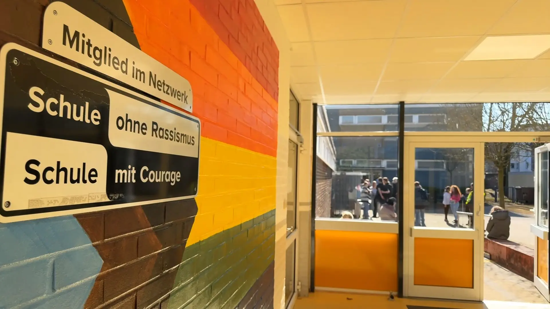 Flur mit einer bunten Wand an der ein Schild hängt "Mitglied im Netzwerk Schule ohne Rassismus – Schule mit Courage". Durch die Tür am Ende des Flurs sieht man auf dem Jugendliche.