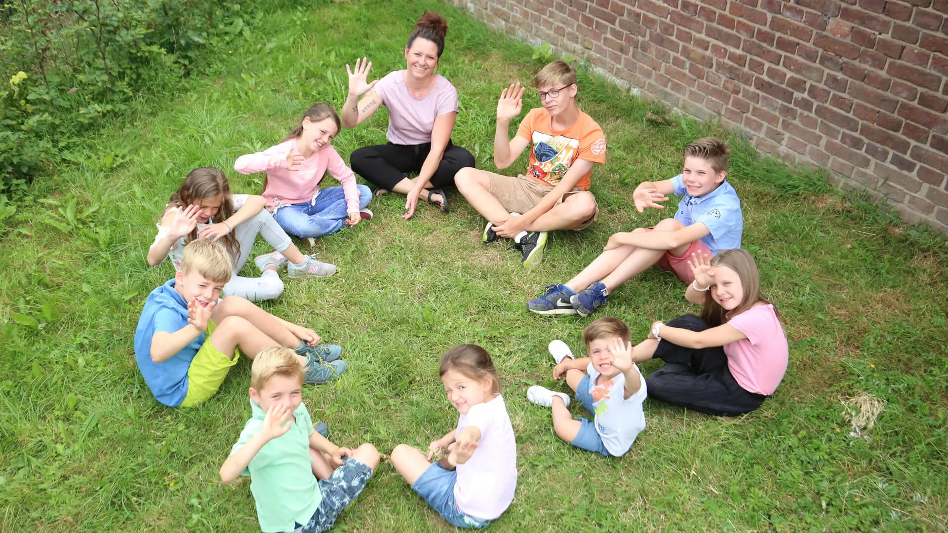 Kinder sitzen mit Erzieherin im Kreis auf einer Wiese zusammen