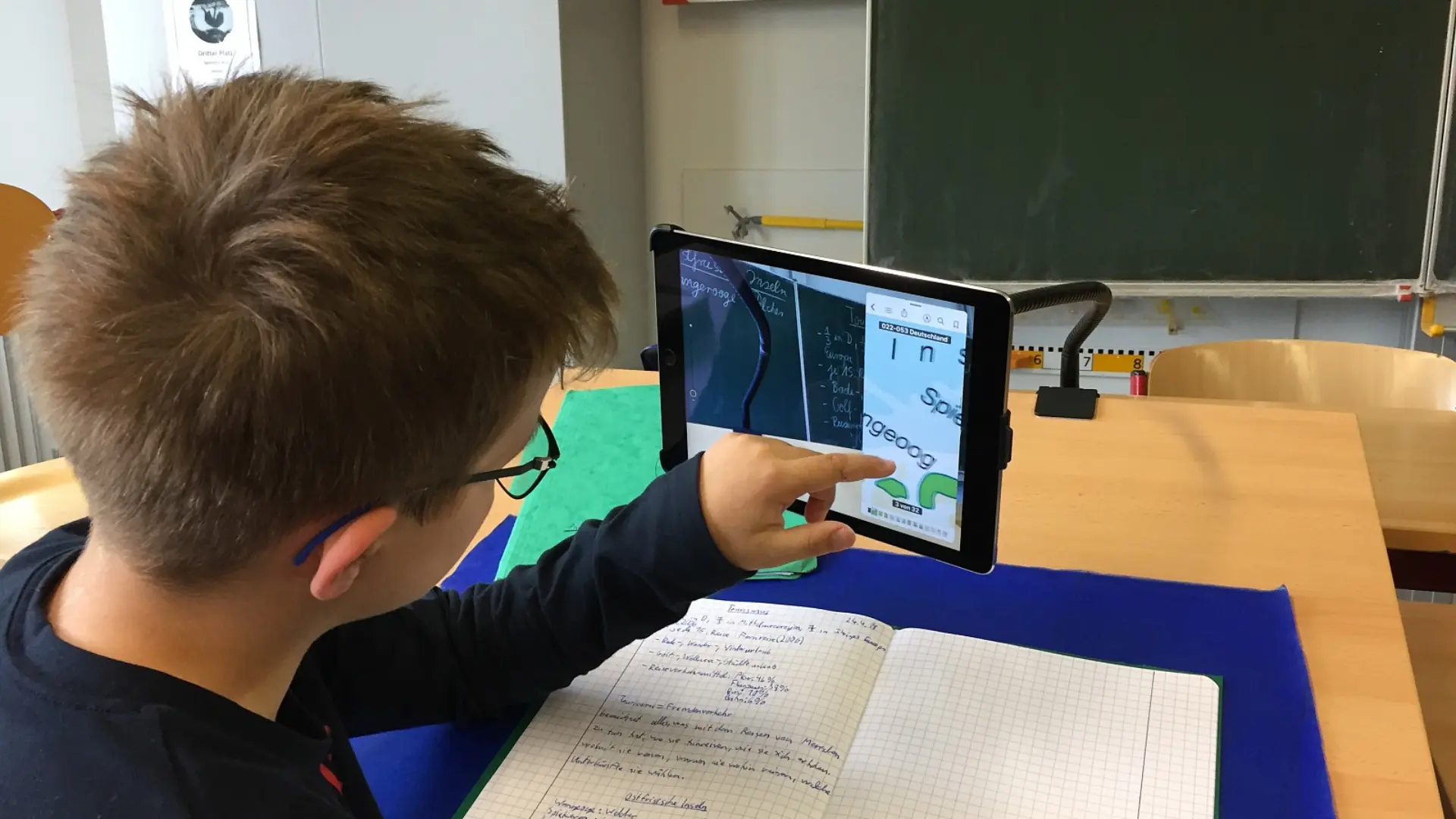 Ein Schüler mit Brille von hinten links gesehen, der an einem Schreibtisch konzentriert mit einem iPad arbeitet. Das Tablet ist an einer stabilen, biegsamen Halterung befestigt. Unter dem iPad liegt ein aufgeschlagenes Schulheft.