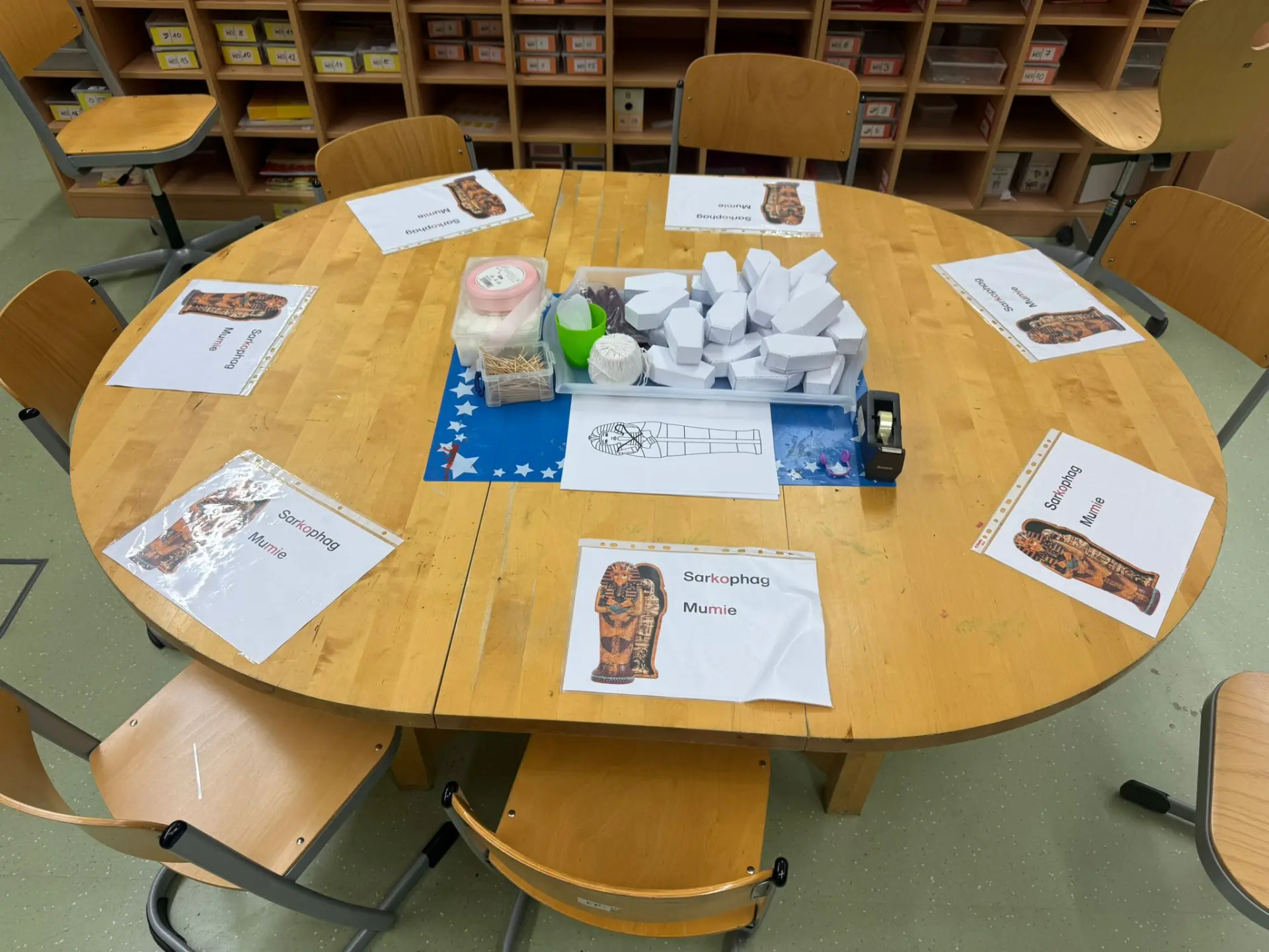 Ein runder Holztisch in einem Klassenzimmer, vorbereitet für ein Projekt zum Thema Ägypten. Auf dem Tisch liegen kreisförmig angeordnete Arbeitsblätter mit der Aufschrift Sarkophag Mumie und entsprechenden Abbildungen. In der Tischmitte steht eine Materialschale mit vielen kleinen Särgen aus Papier, Schnur, Watte und Zahnstochern.