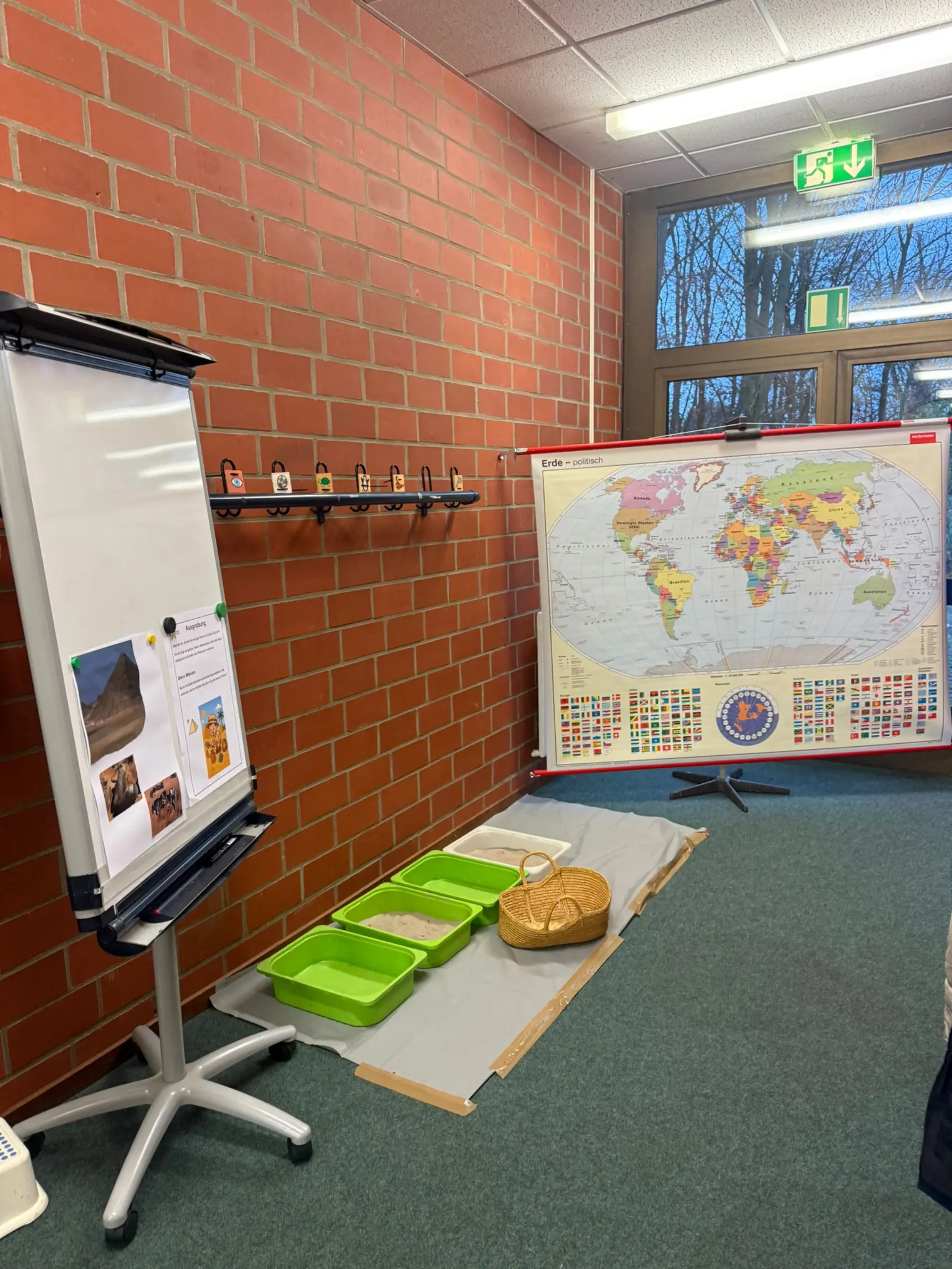 Ein Flurbereich der Schule, der für ein Projekt vorbereitet wurde. Rechts steht eine große Weltkarte auf einem Ständer. Davor sind drei Kunststoffwannen mit Sand, eine Wanne und ein geflochtener Korb aufgestellt. Links im Vordergrund befindet sich ein Flipchart mit Fotos von Pyramiden und Ausgrabungen sowie einem Infoblatt.