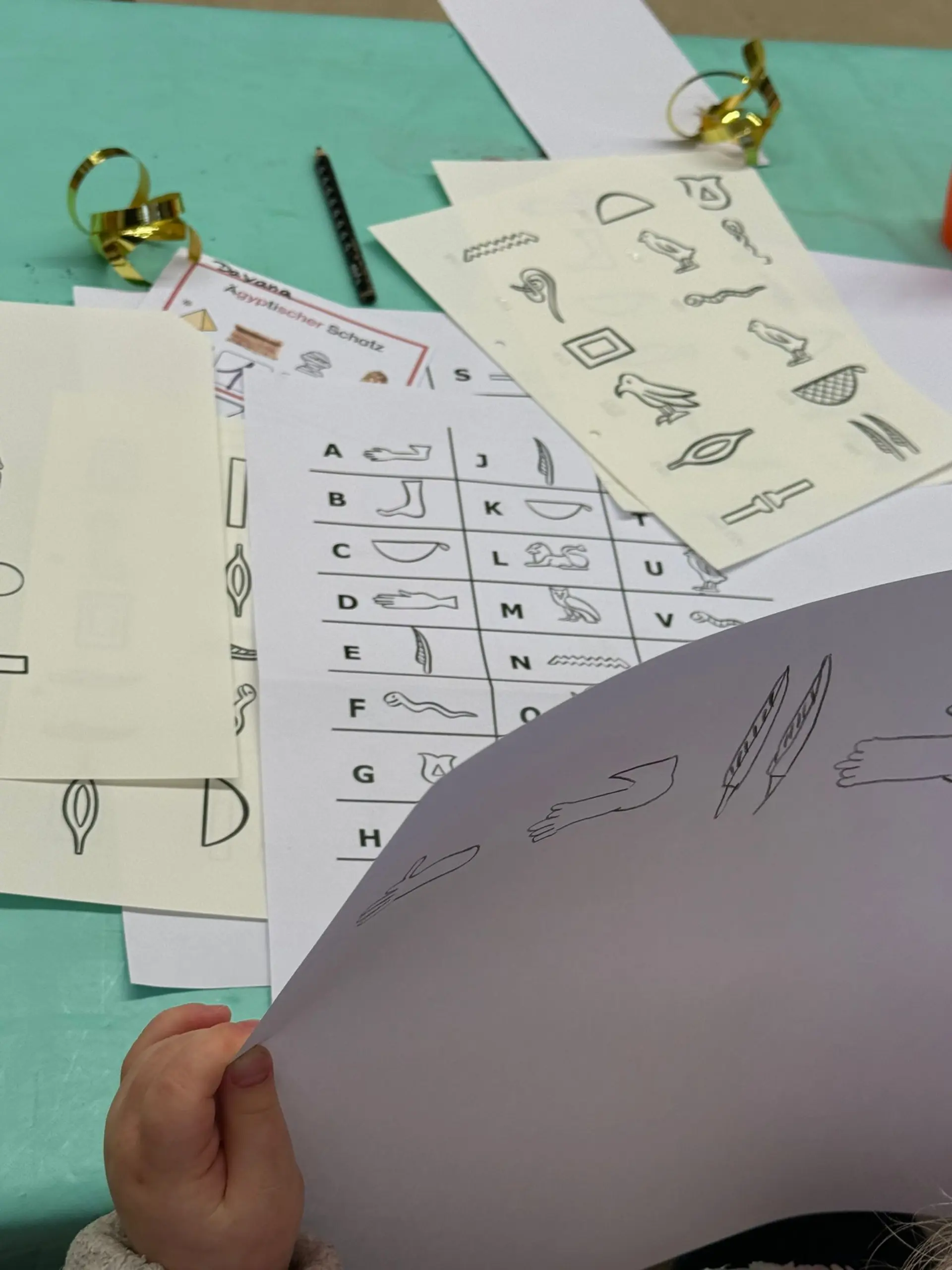 Nahaufnahme von Kinderhänden, die ein Blatt Papier mit selbst gezeichneten Hieroglyphen halten. Auf dem Tisch vor dem Kind liegen weitere Arbeitsblätter, darunter eine Übersicht, die lateinische Buchstaben (A bis V) den entsprechenden ägyptischen Symbolen zuordnet.