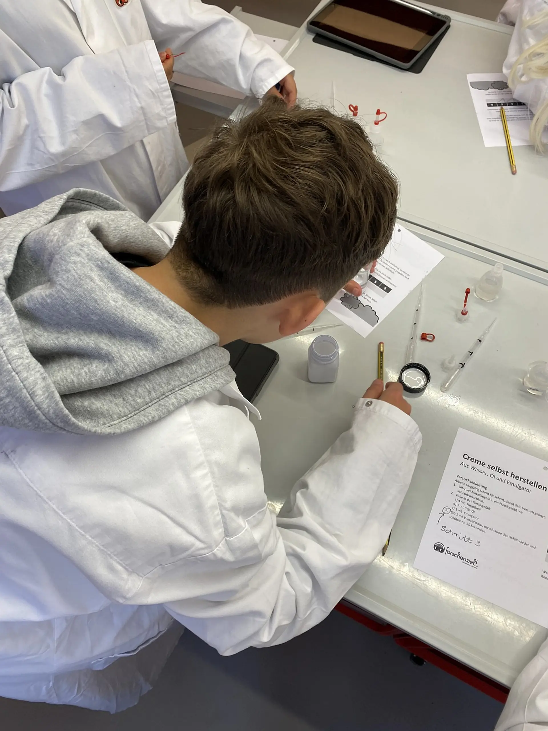 Nahaufnahme von oben eines Schülers im Laborkittel, der konzentriert an einem Experiment arbeitet. Auf dem Labortisch liegen eine detaillierte Anleitung mit dem Titel „Creme selbst herstellen“, Pipetten, kleine Behälter und ein Stift.