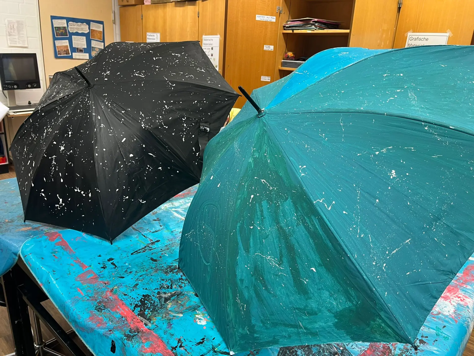 Zwei aufgespannte Regenschirme stehen auf einem mit Schutzfolie abgedeckten Arbeitstisch in einem Kunstraum. Ein Schirm ist tiefschwarz, der andere dunkelgrün grundiert; beide sind mit feinen weißen Farbspritzern überzogen.