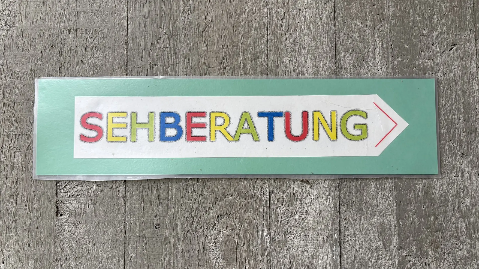 Ein rechteckiges, laminiertes Hinweisschild an einer Wand, das einen Pfeil nach rechts hat. Im Pfeil steht in bunten Großbuchstaben das Wort Sehberatung.