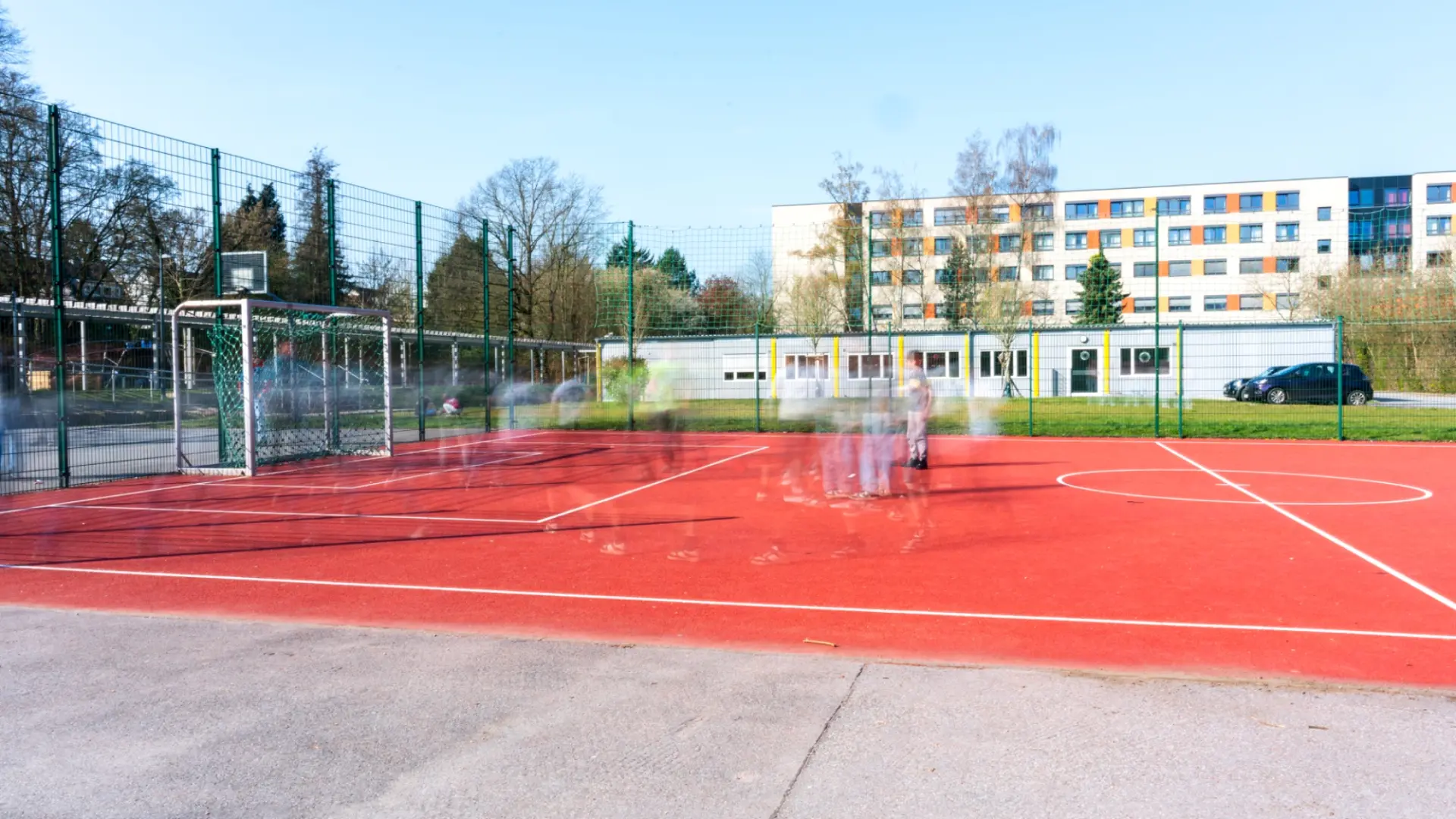 Ein eingezäunter Kleinfeld-Sportplatz mit leuchtend rotem Gummibelag und weißen Spielfeldmarkierungen. Im Vordergrund ist ein Fußballtor mit grünem Netz zu sehen. Durch eine Langzeitbelichtung erscheinen Kinder beim Spielen als schemenhafte, verschwommene Bewegungsspuren.
