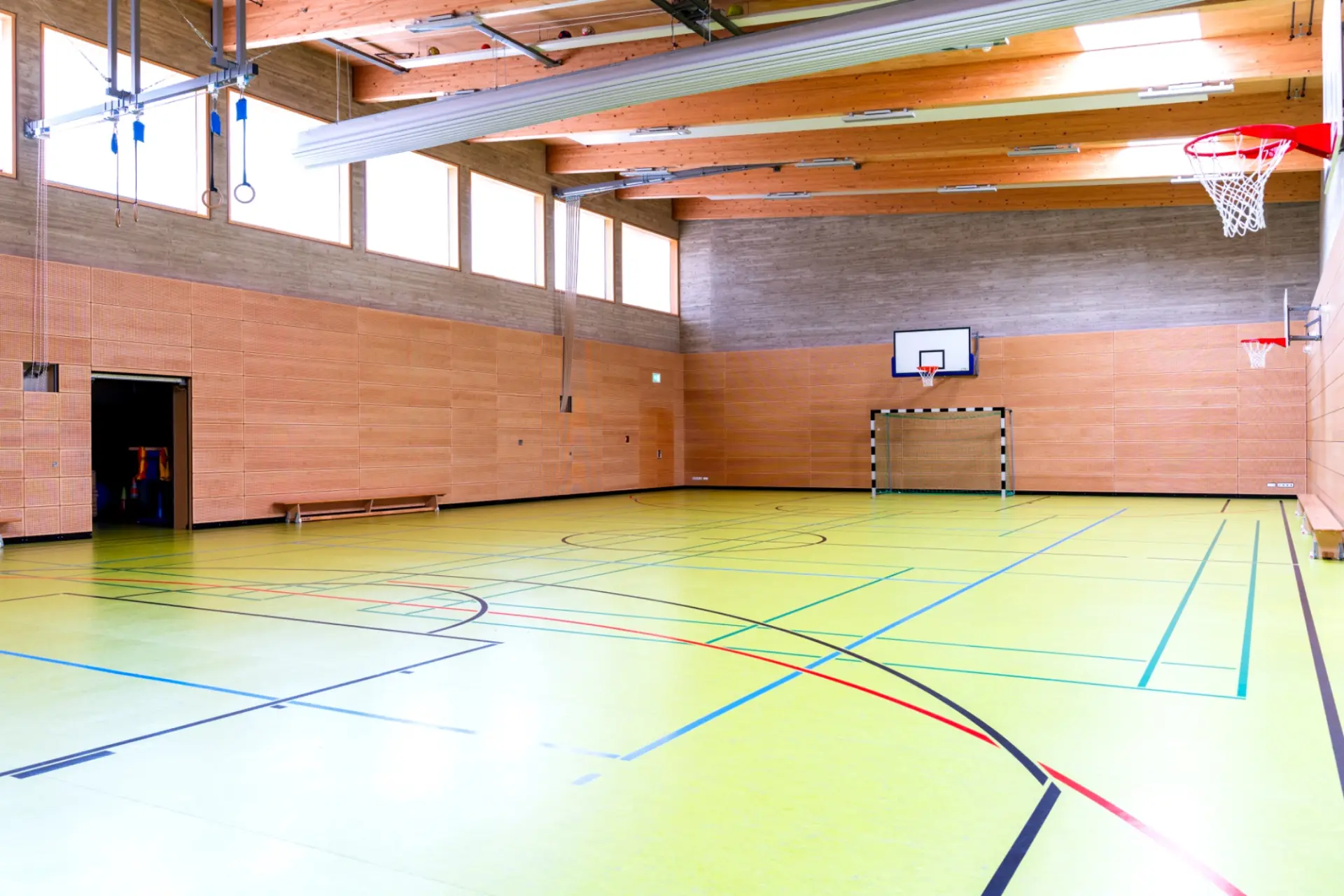 Eine moderne, helle Mehrzweck-Sporthalle mit lindgrünem Boden und verschiedenen Spielfeldmarkierungen. Die Wände sind im unteren Bereich mit hellem Holz getäfelt, während der obere Teil aus Sichtbeton besteht. Große Fensterfronten lassen natürliches Licht herein.