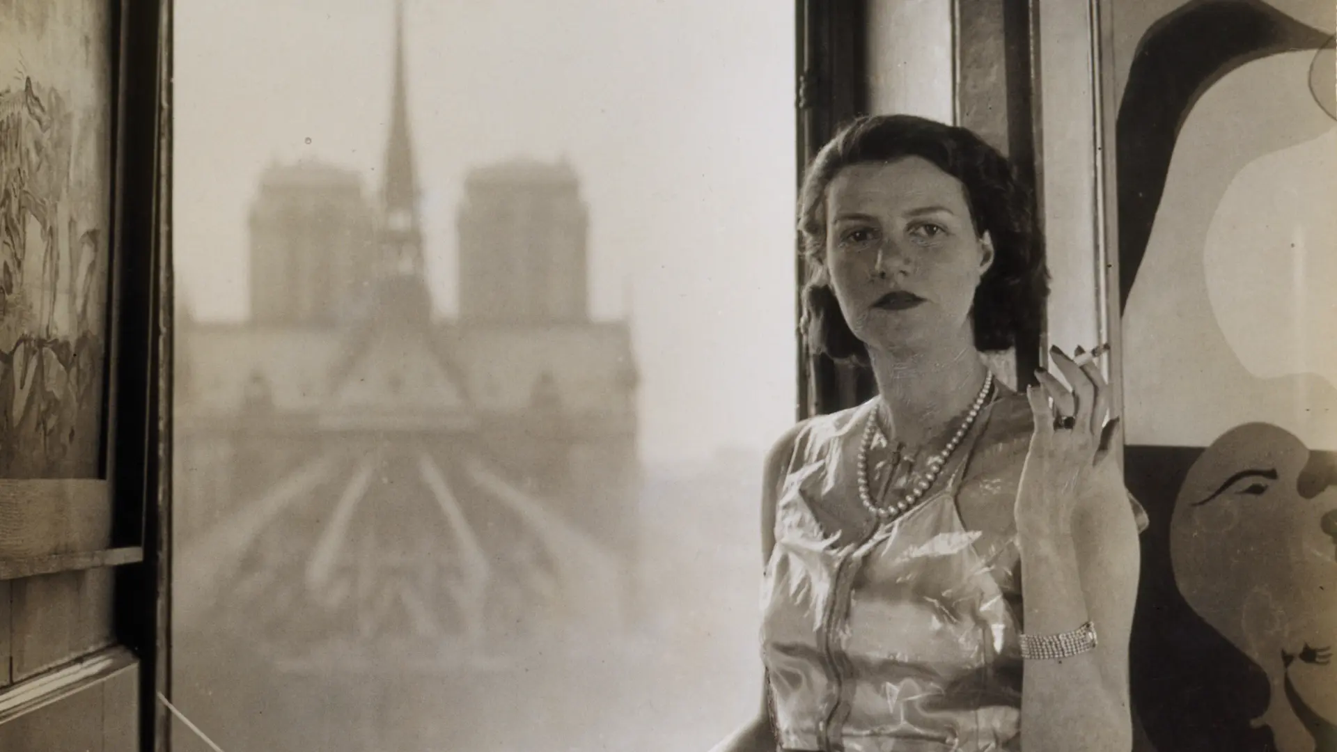 Eine schwarz-weiße Fotografie einer Frau im eleganten Kleid, die vor einem geöffneten Fenster steht.