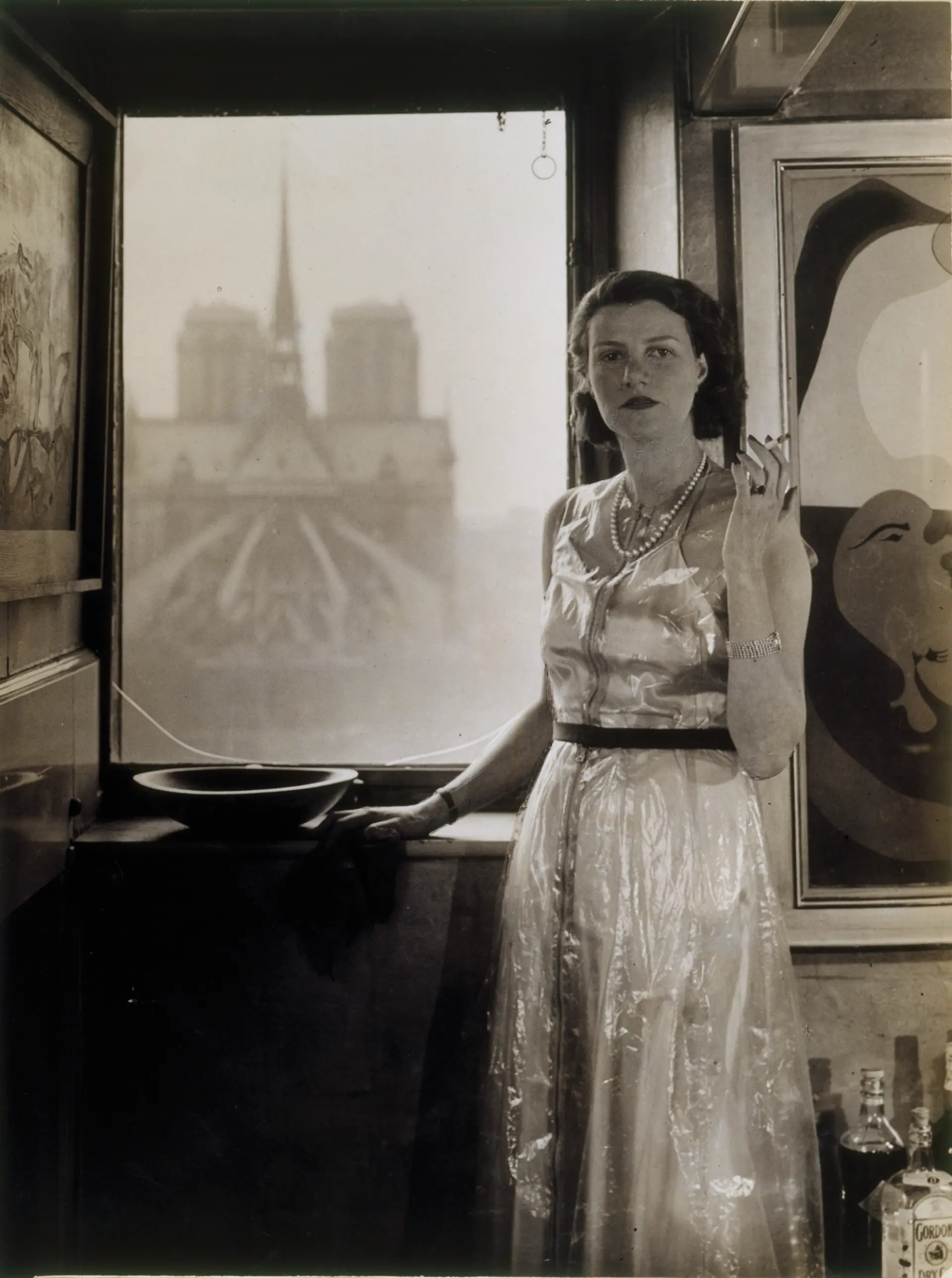 Eine schwarz-weiße Fotografie einer Frau im eleganten Kleid, die vor einem geöffneten Fenster steht.