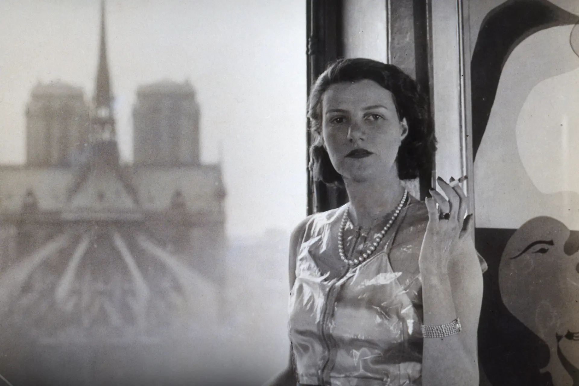 Eine schwarz-weiße Fotografie einer Frau im eleganten Kleid, die vor einem geöffneten Fenster steht.