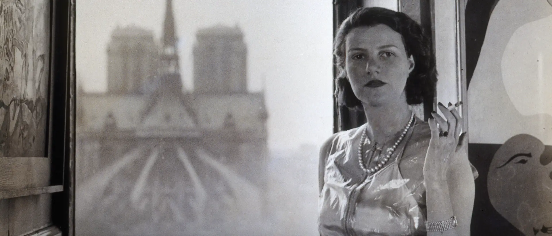 Eine schwarz-weiße Fotografie einer Frau im eleganten Kleid, die vor einem geöffneten Fenster steht.