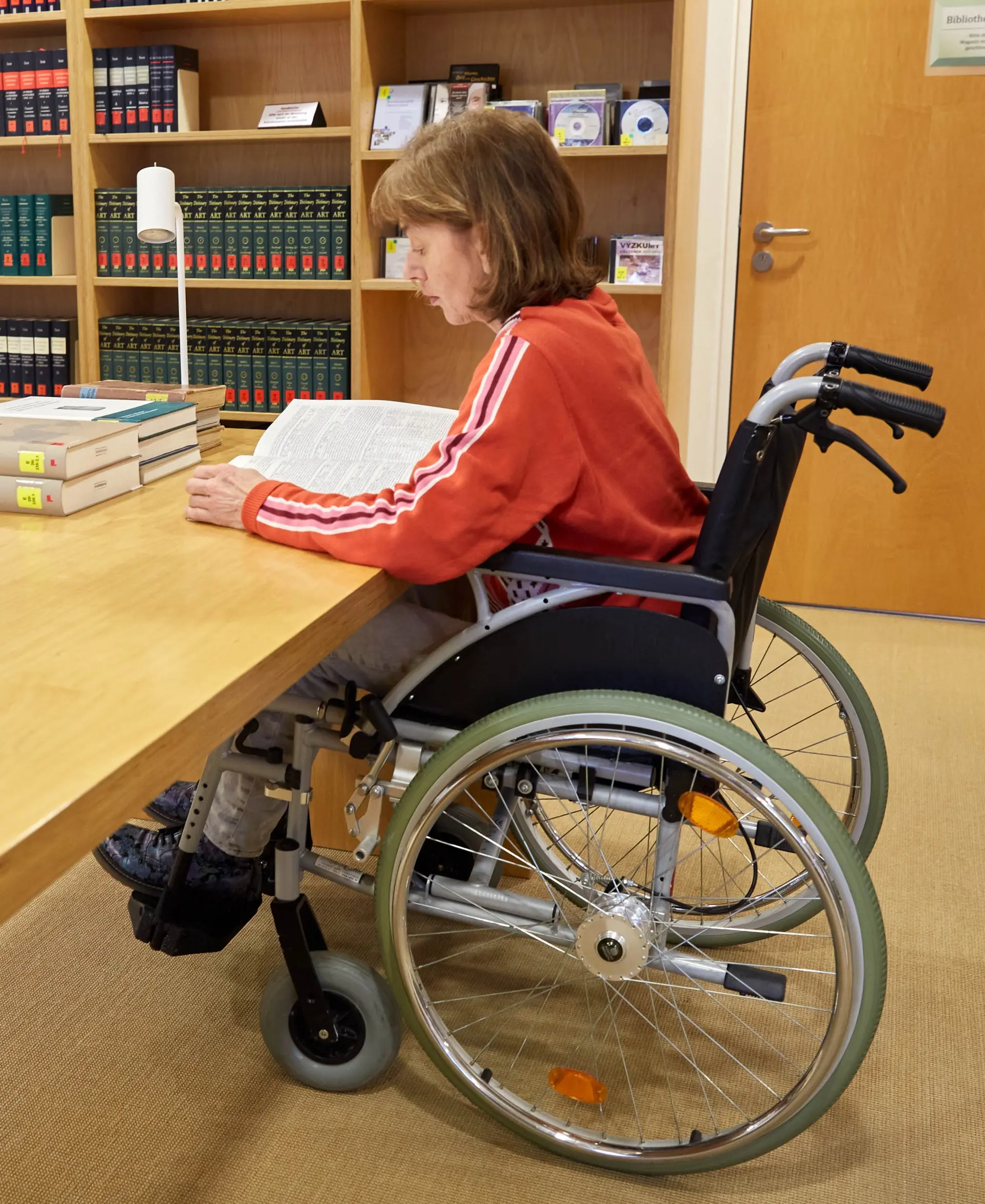 Eine Frau in Rollstuhl liest ein Buch an einem mit dem Stuhl unterfahrbaren Schreibtisch.