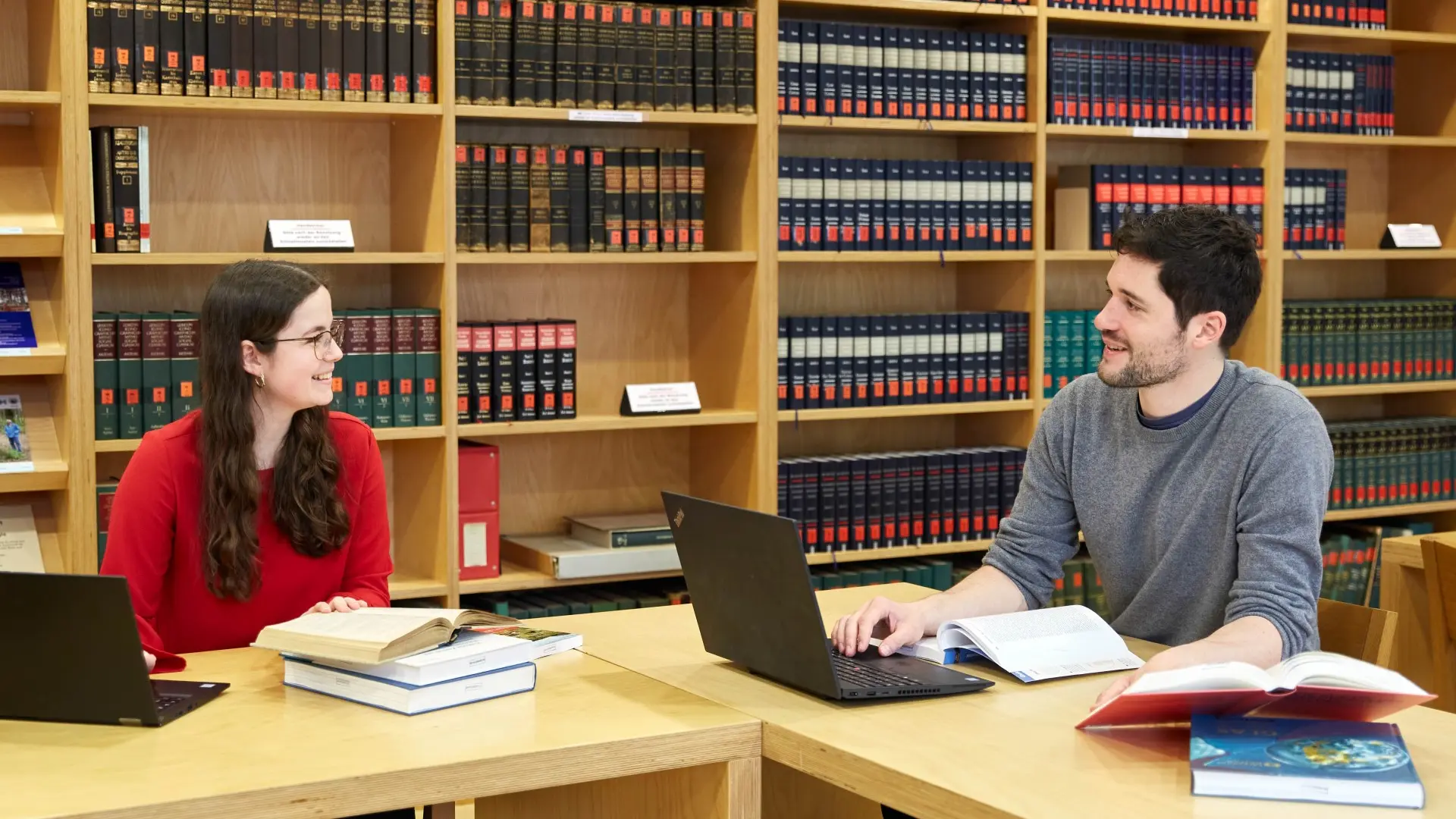 Zwei Menschen an Tischen mit Laptops und Büchern lächeln sich an.