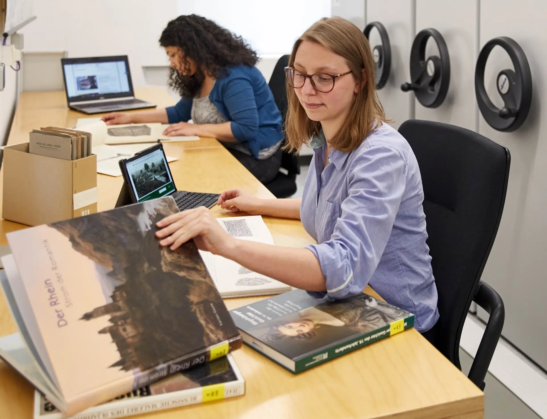 Zwei Studierende recherchieren mit Büchern und Computern an einem Arbeitsplatz.