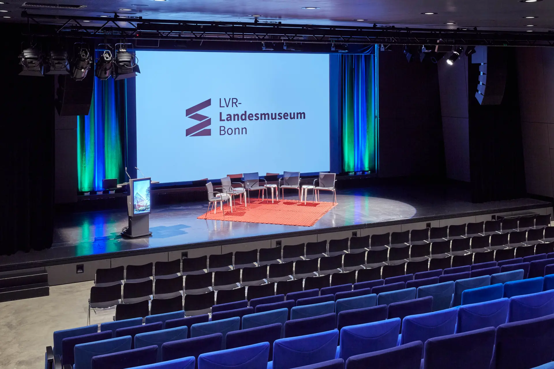 Ein Blick im Veranstaltungssaal des LVR-Landesmuseums Bonn vom bestuhlten Publikumsbereich auf die Bühne. Auf der Bühne stehen ein Rednerpult sowie fünf Stühle auf einem Teppich vor einem Bild des Landesmuseums im Hintergrund.