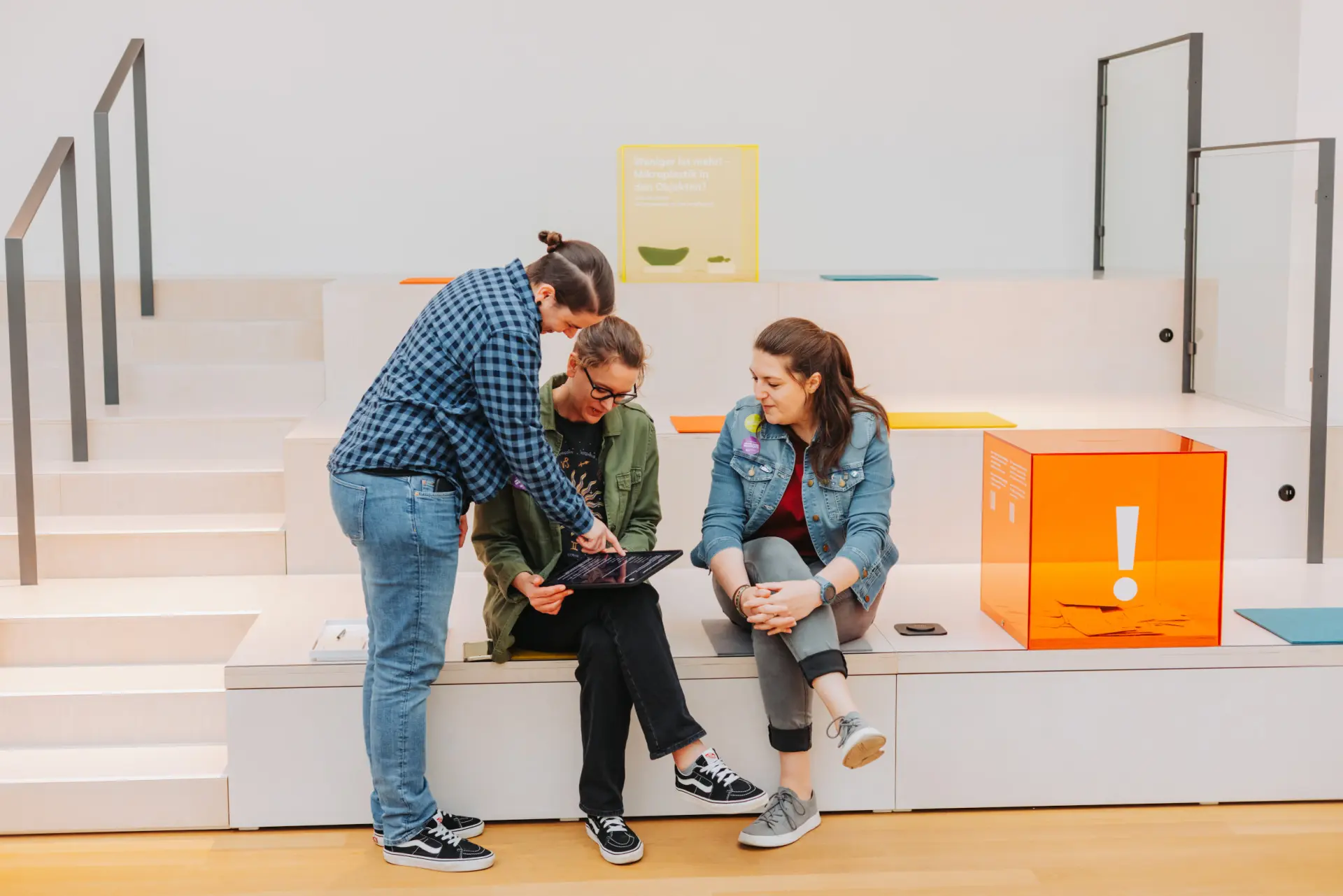 Drei Personen sitzen in einem Museumsbereich aus hellem Holz mit bunten Farbakzenten, dem "Museum der Zukunft".