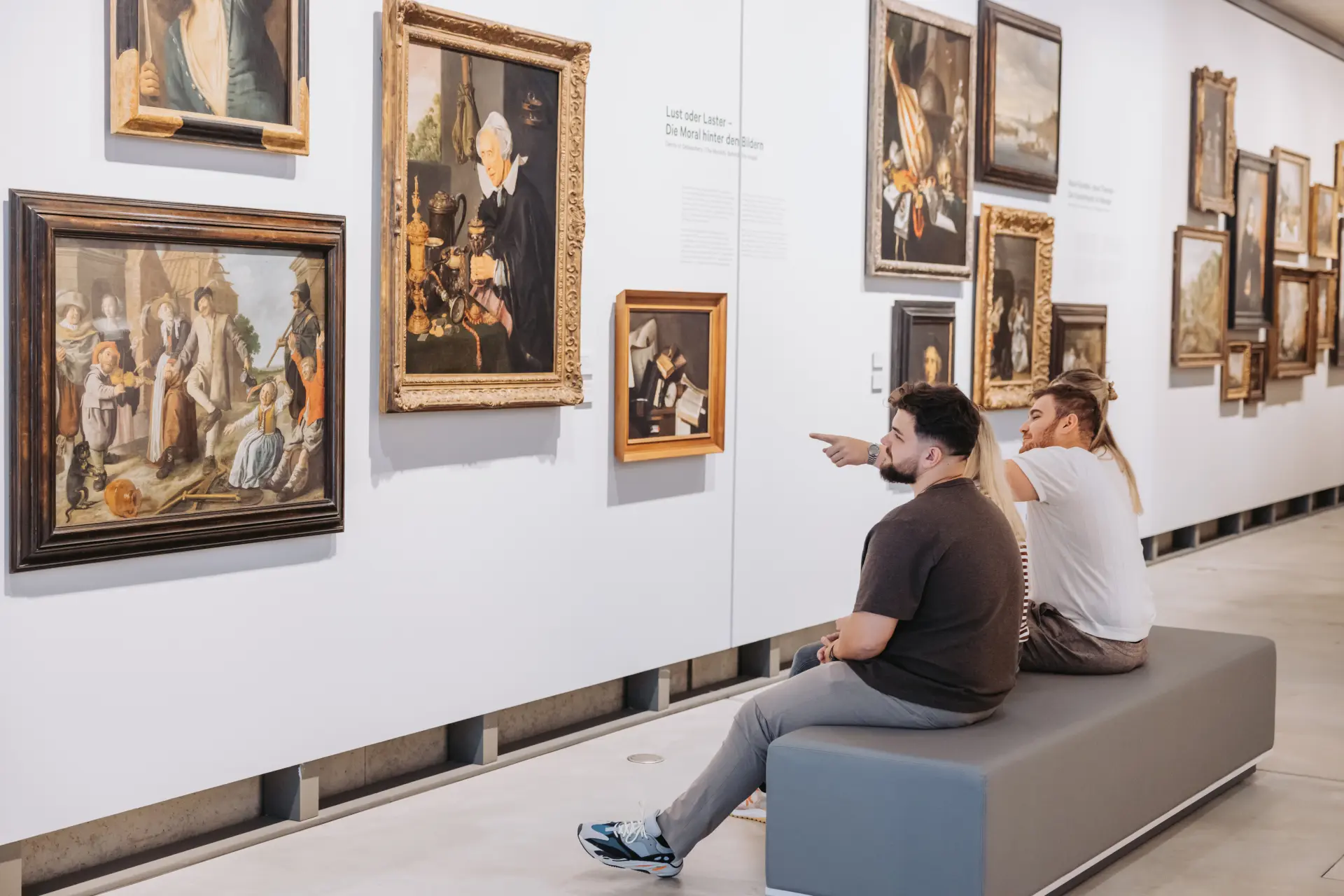 Eine Gruppe junger Erwachsener sitzen in der Galerie auf einer Bank und schauen sich Gemälde an.