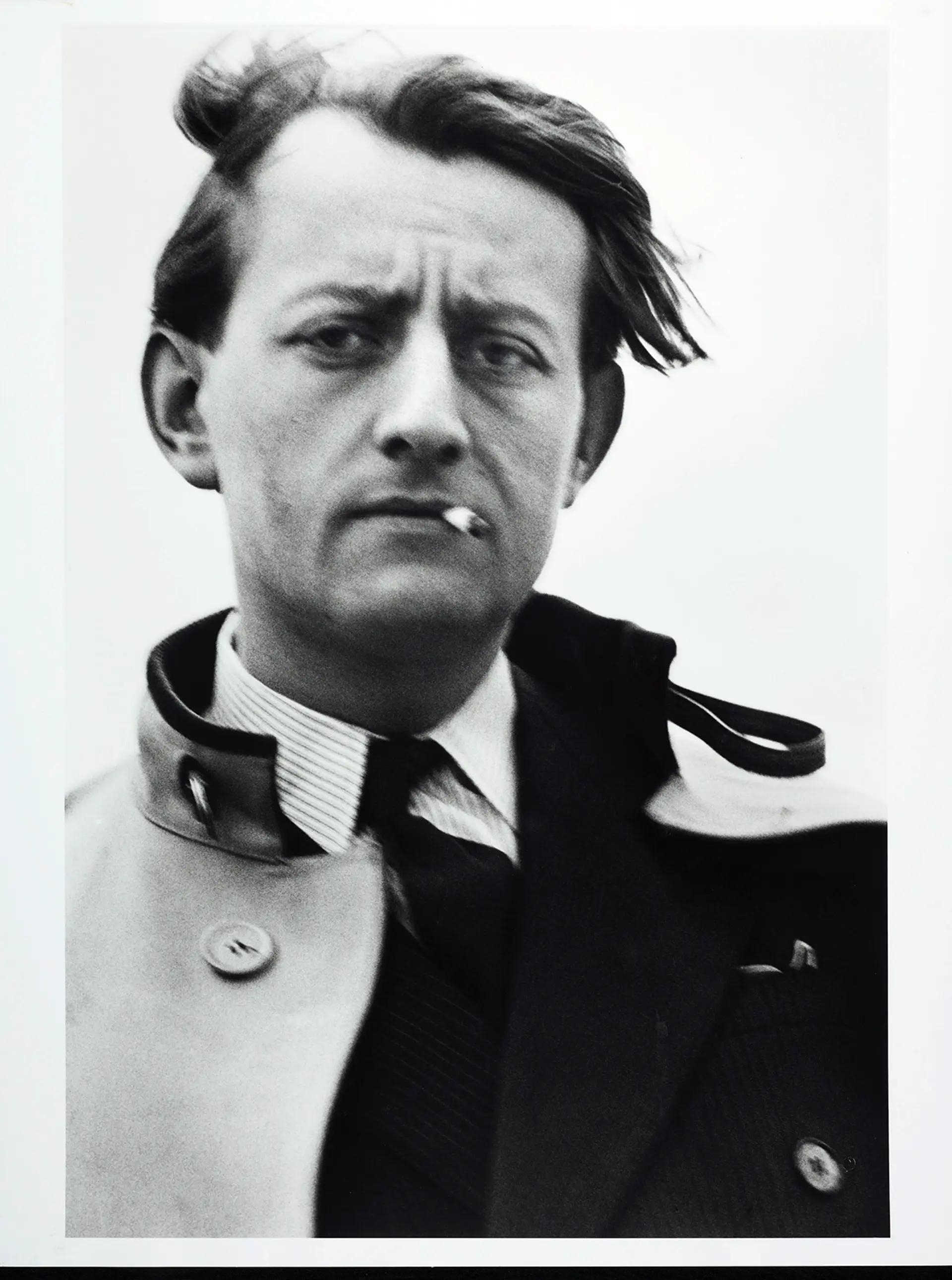 Ein Mann in Anzug und im Wind flatternder Jacke. Er schaut ernst und hat eine Zigarette im Mundwinkel.