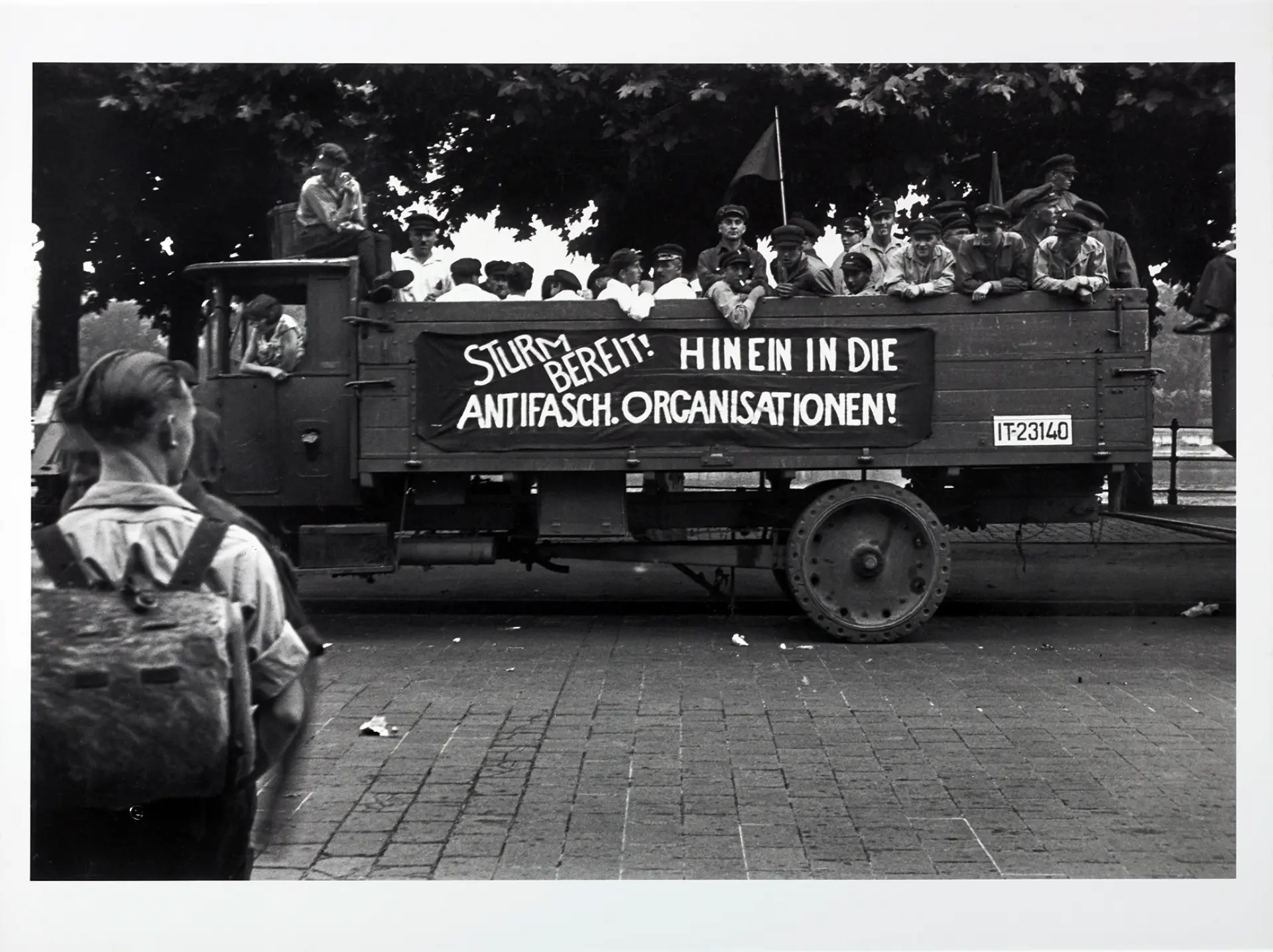 Ein Lastwagen, auf dem eine Reihe uniformierter Männer stehen. Auf der Seite des Lastwagens hängt ein Banner mit der Aufschrift "Sturm bereit! Hinein in die Antifaschistischen Organisationen!"