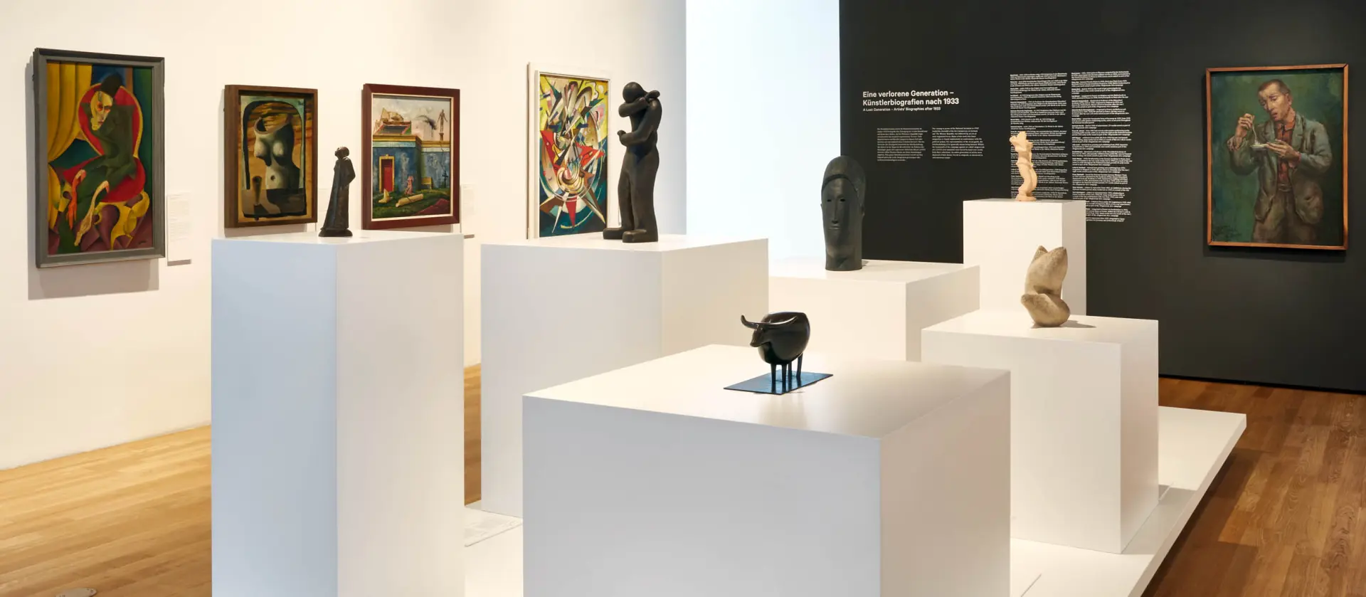 Im Ausstellungsbereich Moderne stehen viele unterschiedliche Skulpturen auf verschiedenhohen weißen Sockeln. Im Hintergrund hängen Gemälde mit abstrahierten Figuren an der Wand.  