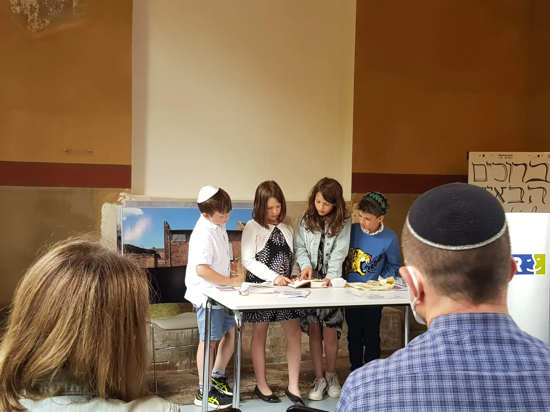 Vier Kinder stehen in der Synagoge vor der Tora-Nische und lesen aus einem Buch, das auf einem Tisch vor ihnen liegt.