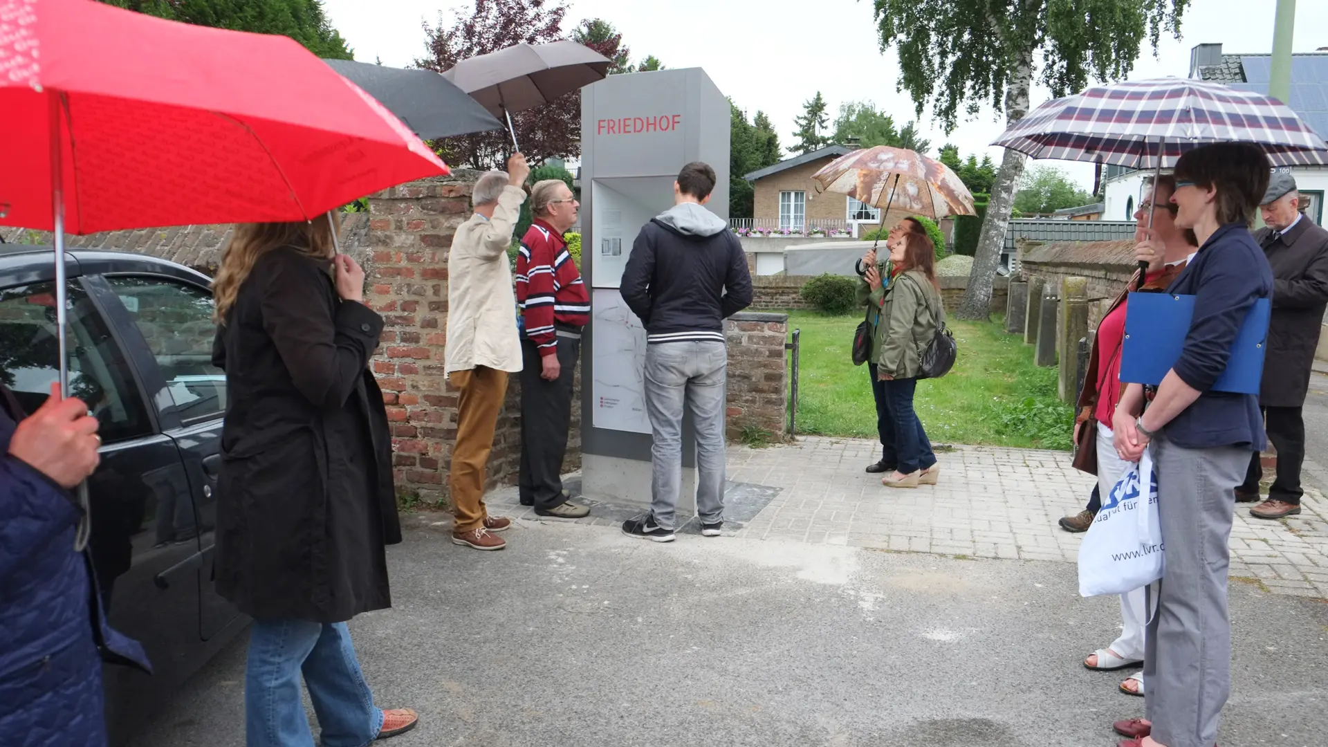 Mehrere Personen mit Regenschirmen stehen am Eingang zum jüdischen Friedhof. Sie blicken auf eine graue Stele, die die Inschrift "Friedhof" hat.