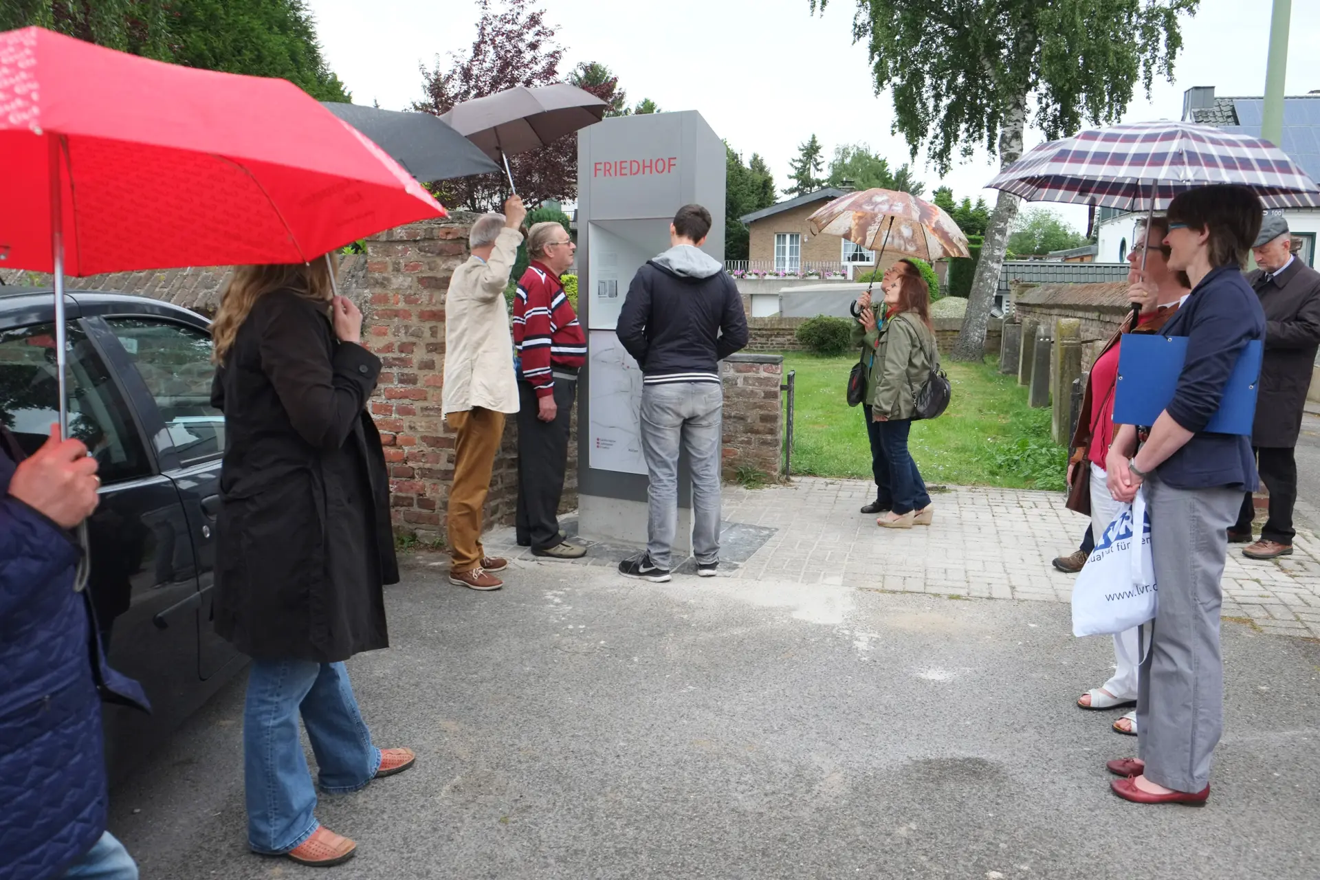 Mehrere Personen mit Regenschirmen stehen am Eingang zum jüdischen Friedhof. Sie blicken auf eine graue Stele, die die Inschrift "Friedhof" hat.