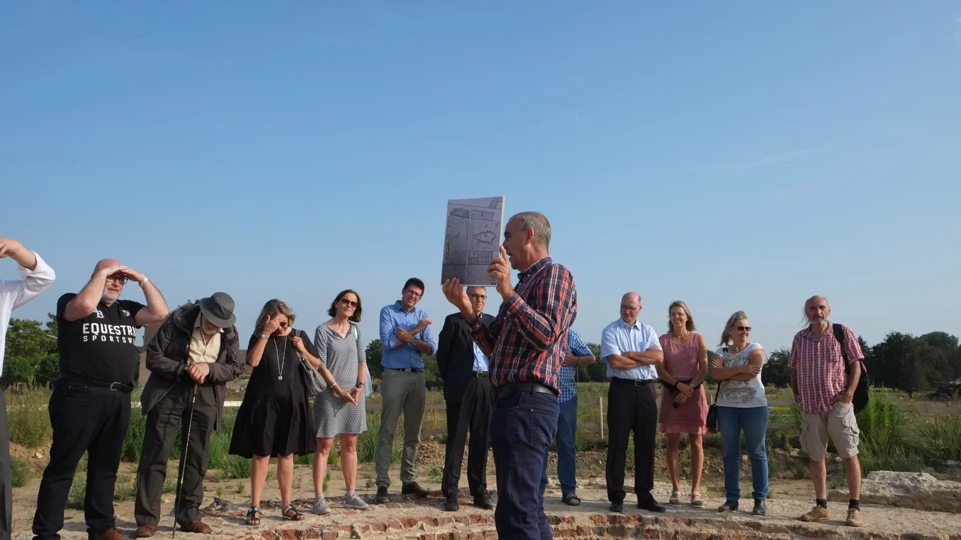 Mehrere Menschen stehen auf freiem Feld an einer Baugrube. Eine Person erklärt etwas.