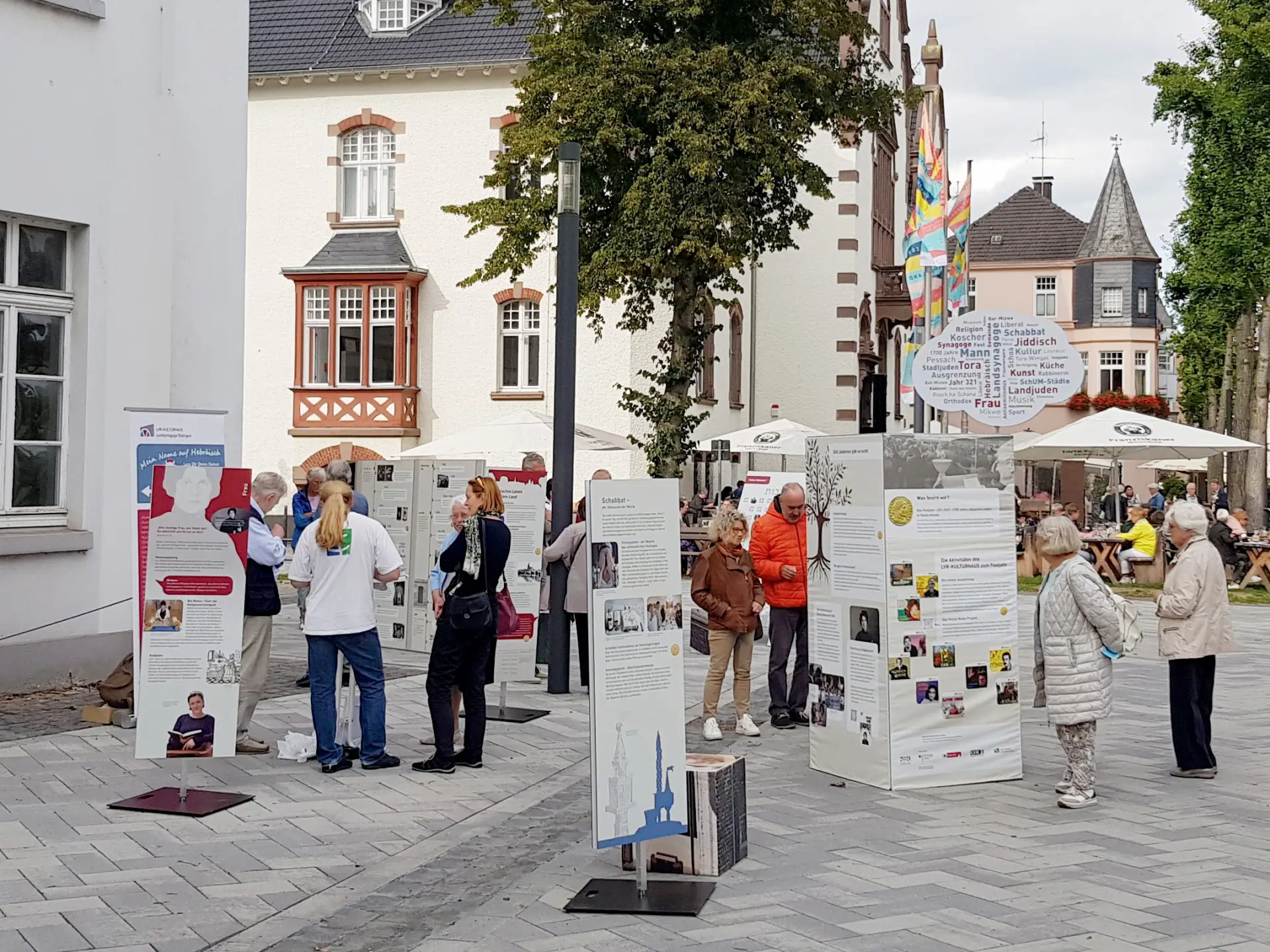 Menschen stehen auf einem Platz umgeben von mittelalterlichen Gebäuden und betrachten die Aufsteller einer Ausstellung.
