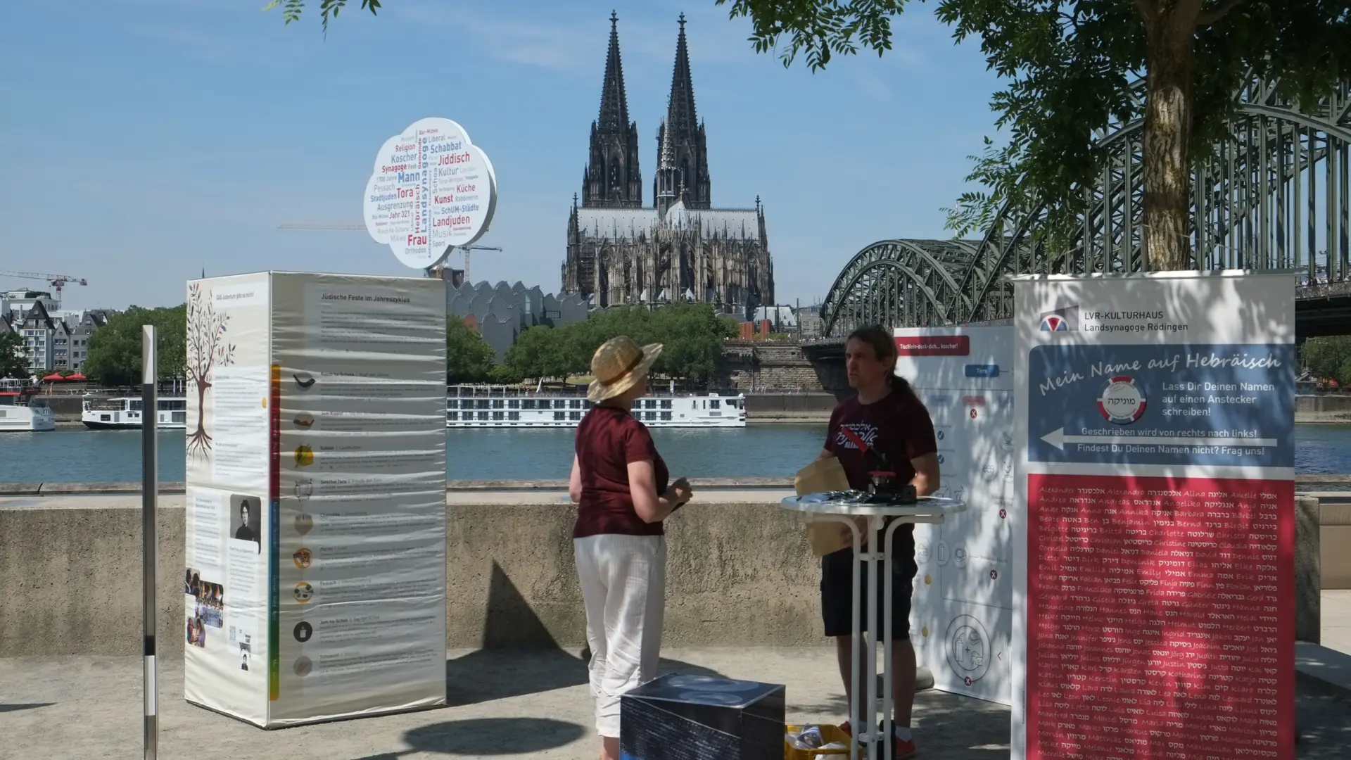 Eine Frau mit Strohhut und ein Mann stehen zwischen Aufstellern einer Ausstellung am Rhein. Im Hintergrund ist die Hohenzollernbrücke und der Dom zu sehen.