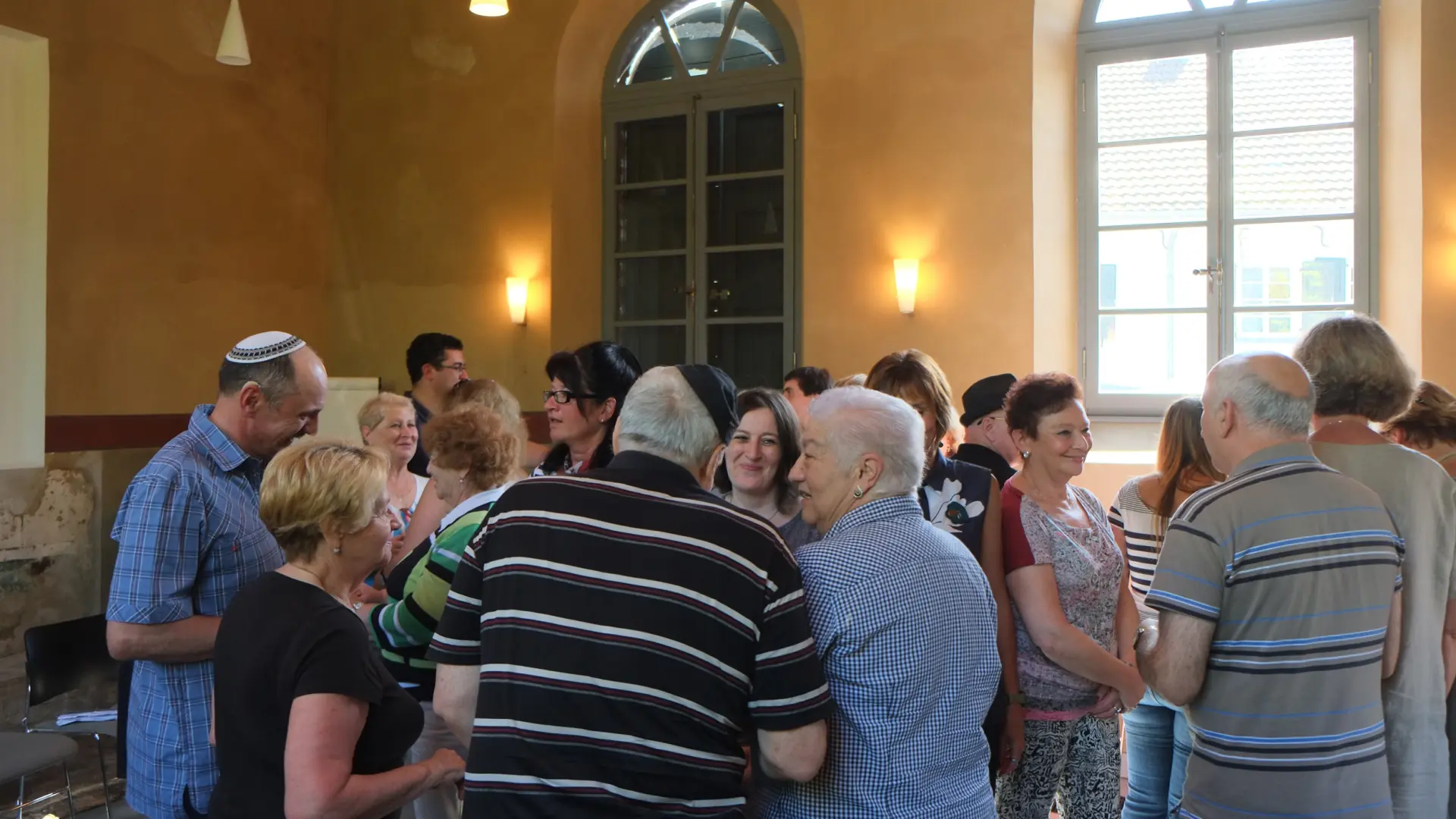 Eine Gruppe von Menschen steht in der Synagoge. Die Menschen unterhalten sich untereinander.