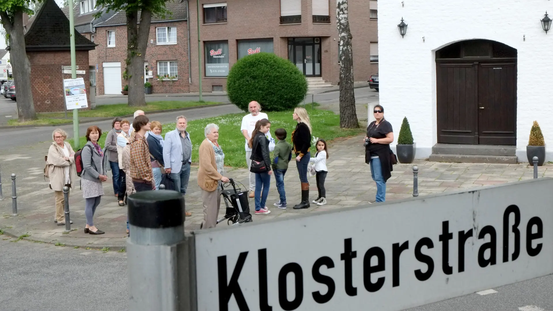 Blick über das Straßenschild "Klostergasse" auf eine Gruppe von Menschen, die an einem geführten Dorfrundgang zu jüdischen Orten im christlichen Dorf teilnehmen.