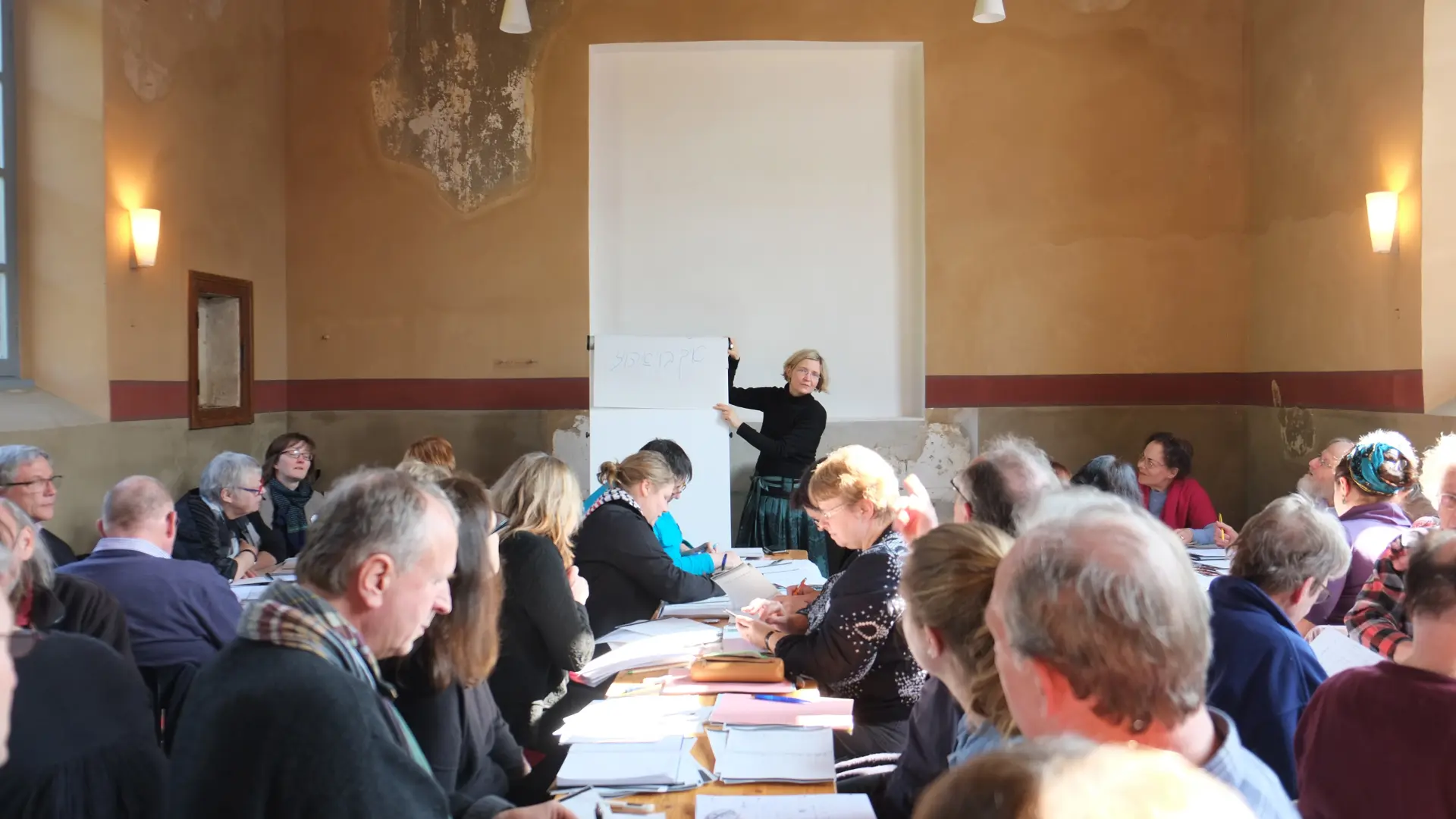 In einem voll besetzten Raum, der Landsynagoge, schaut das Publikum zu einer Frau an einer Schreibtafel. Die Frau verweist erklärend auf die Tafel.