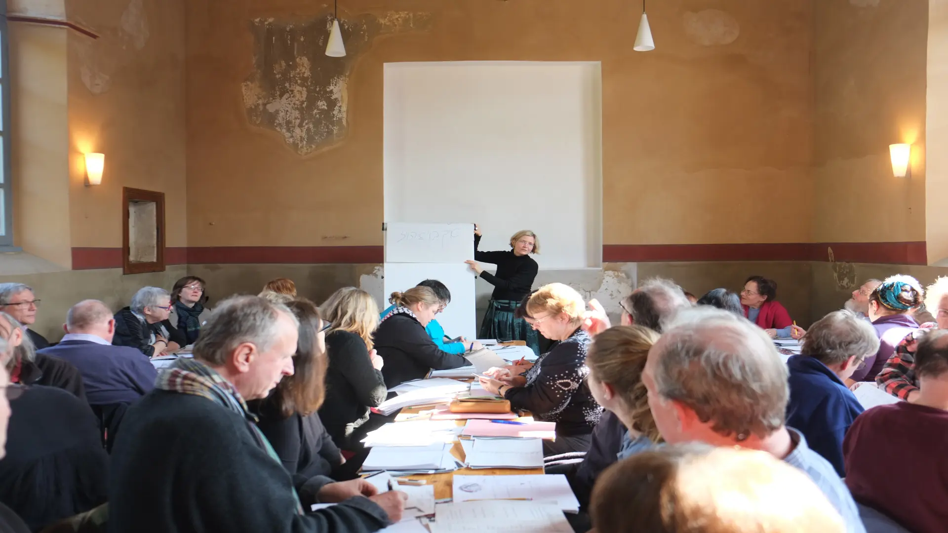 In der Synagoge sitzen mehrere Menschen an Tischen mit Arbeitsmaterial. Vor der Tora-Nische steht eine Frau, die an einer Tafel etwas erklärt.