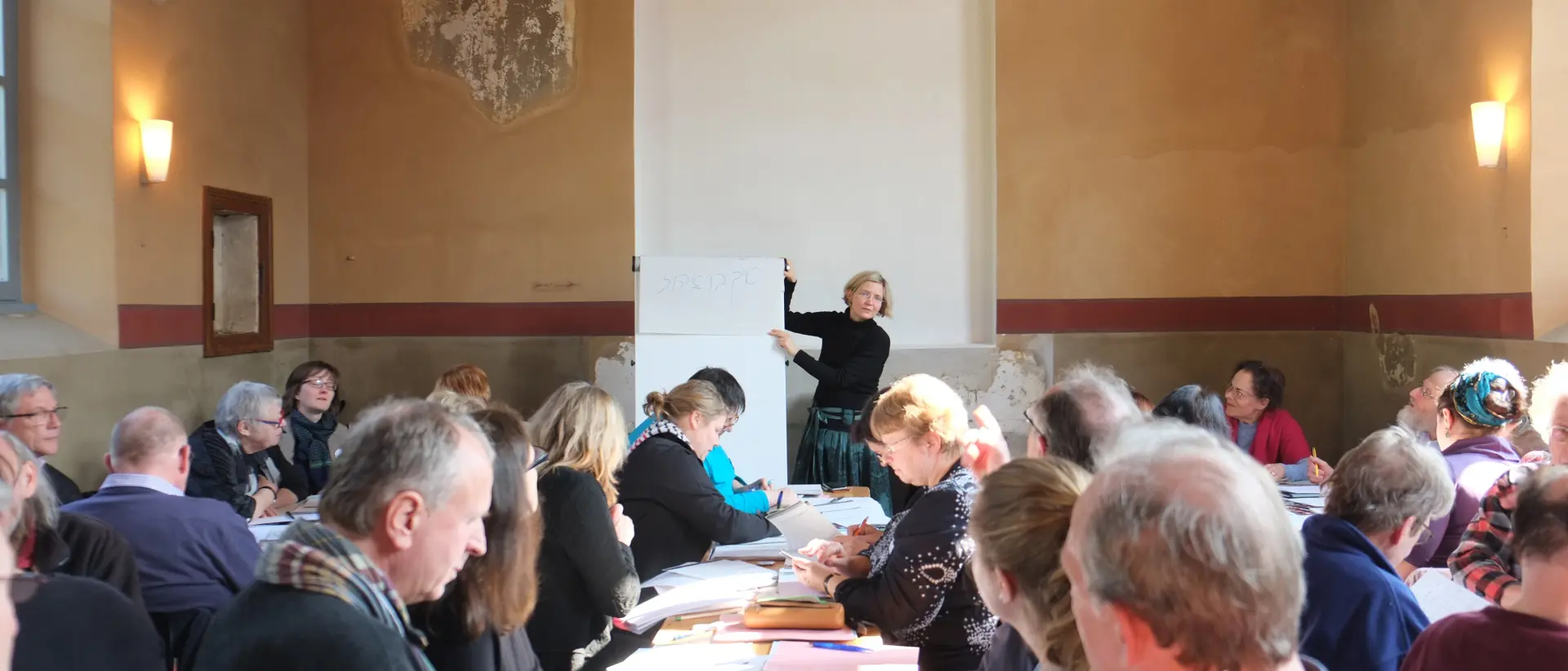 In der Synagoge sitzen mehrere Menschen an Tischen mit Arbeitsmaterial. Vor der Tora-Nische steht eine Frau, die an einer Tafel etwas erklärt.