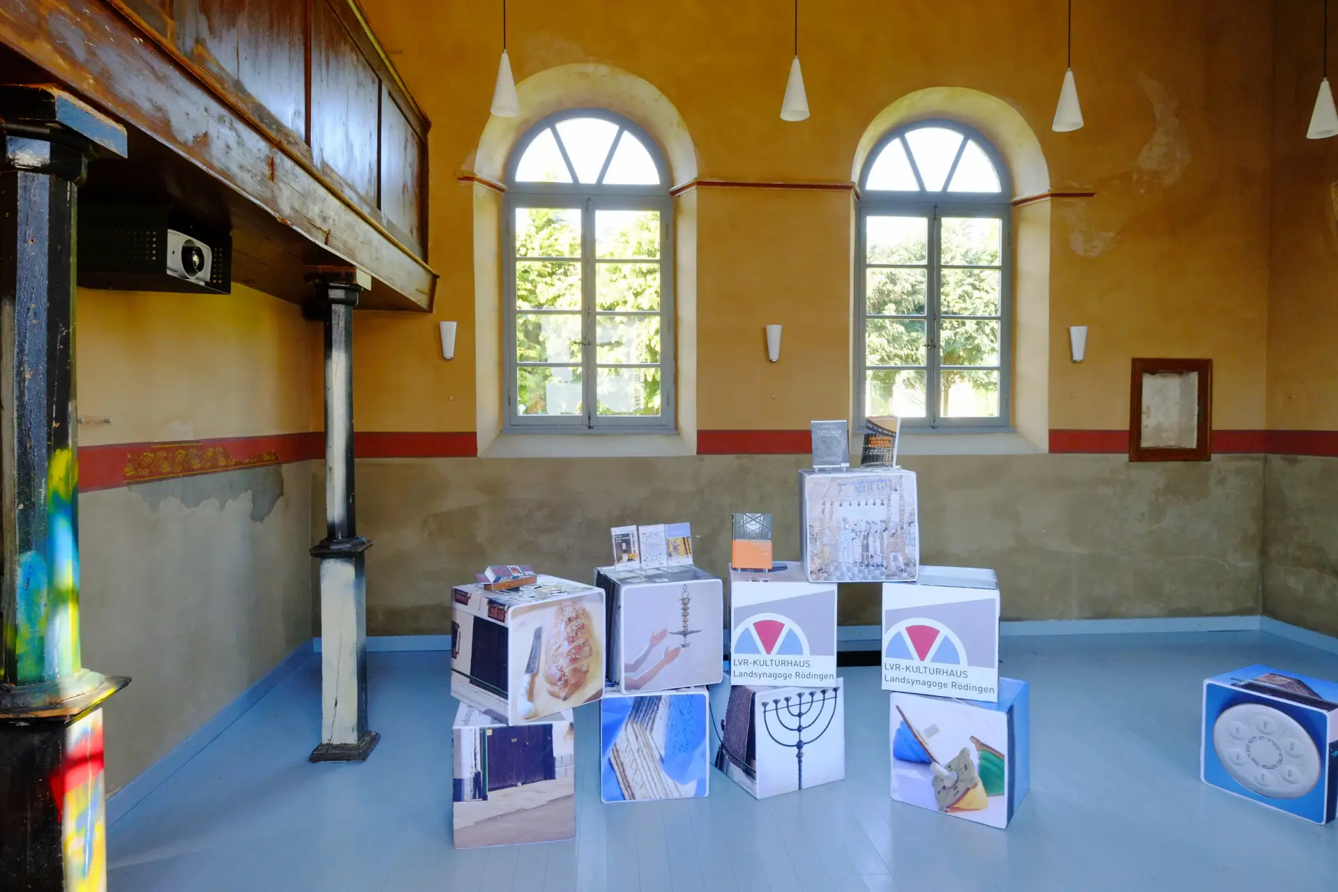 Blick in den Innenraum der Synagoge. Auf der linken Seite sieht man die Frauenempore. Sie ist gestützt von zwei Säulen, die bunte Farbspritzer aufweisen. In der gegenüberliegenden Wand ist ein hochrechteckiges Fensterpaar. Auf dem Holzboden der Synagoge sind Sitzwürfel mit verschiedenen Bildmotiven arrangiert.