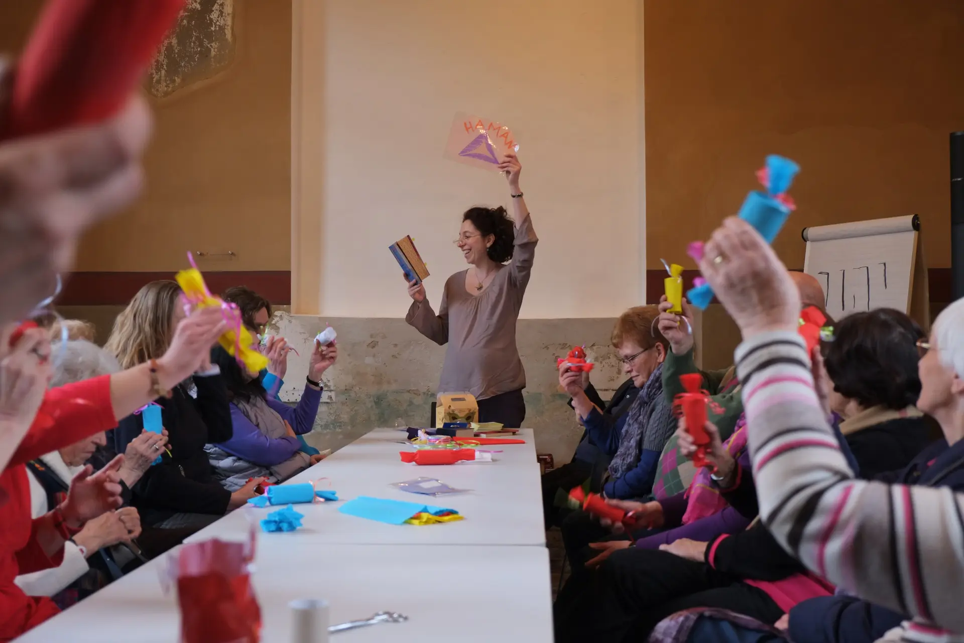 An einem langen Tisch sitzen mehrere Personen mit selbstgebastelten, bunten Rasseln in ihren erhobenen Händen. Am Ende des Tisches steht eine Frau, die aus einem Buch rezitiert und eine Karte emporhält.