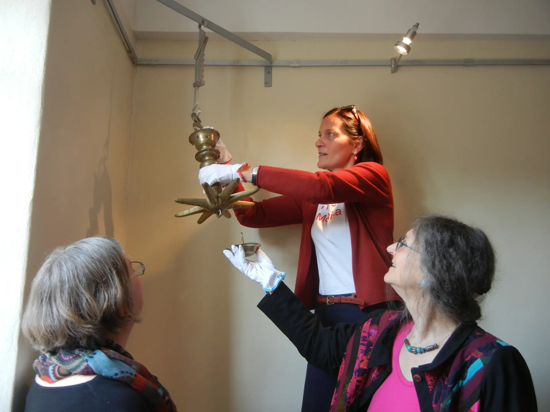 Drei Frauen hängen eine alte Messing-Lampe an eine Aufhängevorrichtung in Ausstellung.