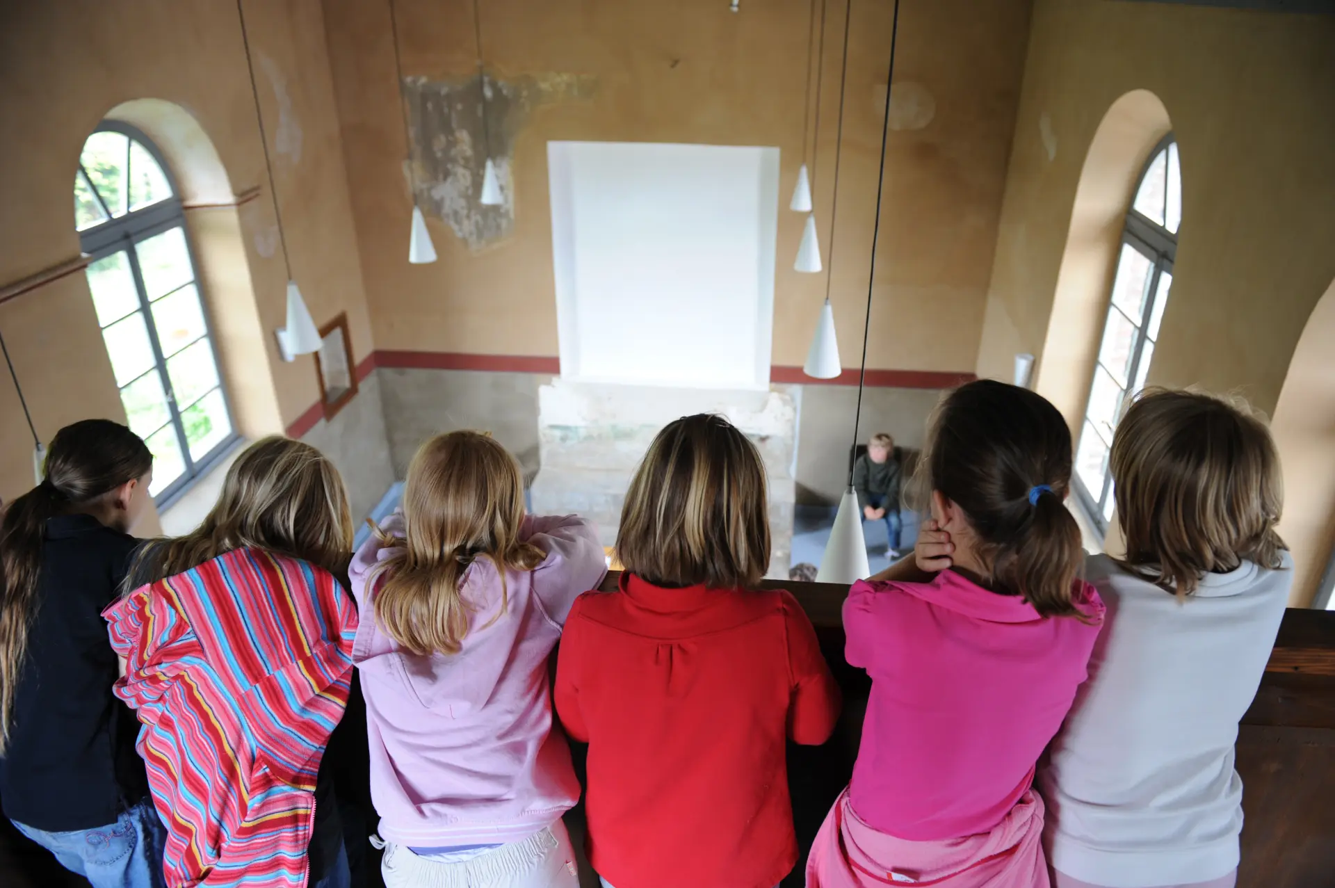 Blick in den Innenraum der Synagoge. Auf der linken Seite sieht man die Frauenempore. Sie ist gestützt von zwei Säulen, die bunte Farbspritzer aufweisen. In der gegenüberliegenden Wand ist ein hochrechteckiges Fensterpaar. Auf dem Holzboden der Synagoge sind Sitzwürfel mit verschiedenen Bildmotiven arrangiert.