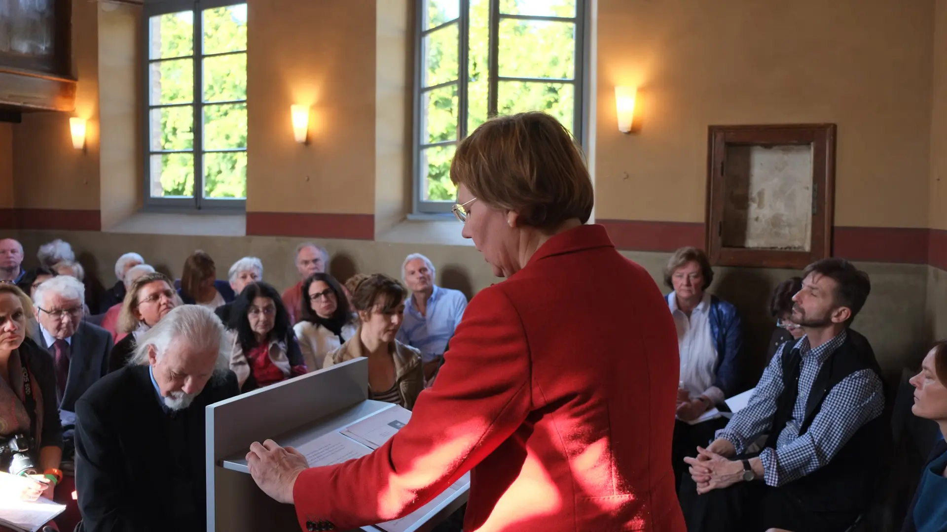 In der Synagoge sitzen mehrere Menschen an verschiedenen Tischen. Ein Mann steht vor ein Roll-up mit dem hebräischen Alef-Bet und deutet mit der Hand auf die abgebildeten Buchstaben.