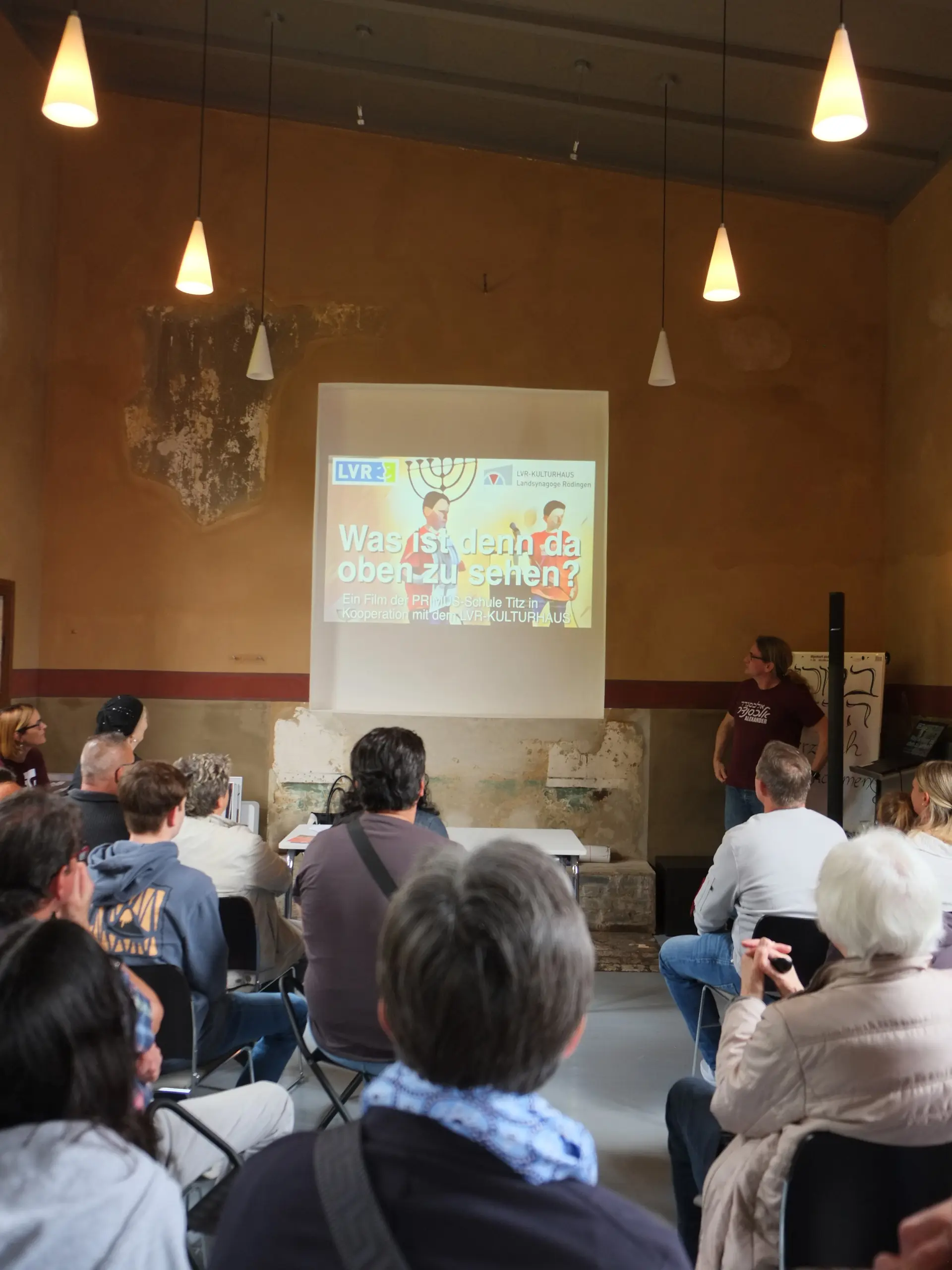 Blick in den Synagogenraum. Menschen sitzen auf Stühlen und schauen auf die gegenüberliegende Wand, auf der das Titelbild des Films "Was ist denn da oben zu sehen?" projiziert ist.