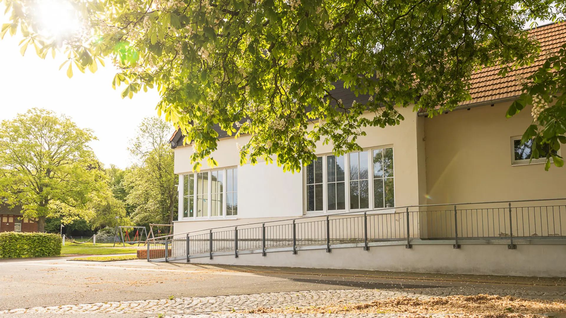 Ein Schulgebäude mit einer Rollstuhlauffahrt aufm dem Schulgelände im Vordergrund, im Hintergrund ist ein Spielplatz zu sehen.