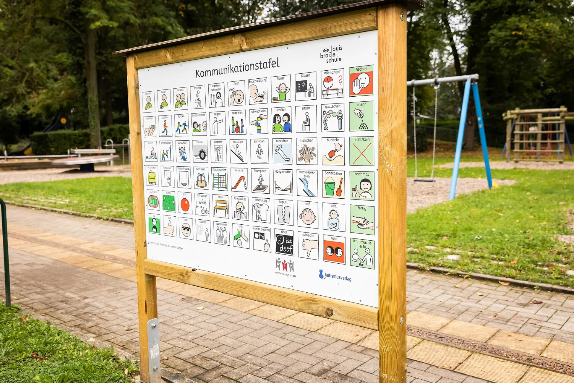 Eine Holztafel mit der Aufschrift "Kommunikationstafel" steht auf einem gepflasterten Weg draußen auf dem Schulgelände. Die Tafel ist mit vielen kleinen Metacom-Piktogrammen versehen, die verschiedene Aktivitäten, Gefühle und Objekte darstellen, um die Kommunikation zu erleichtern. Rechts oben auf der Tafel ist das Logo der "LVR-Louis-Braille-Schule" zu sehen. Im Hintergrund befindet sich ein Spielplatz mit Schaukeln und anderen Geräten, umgeben von Bäumen und Grünflächen.